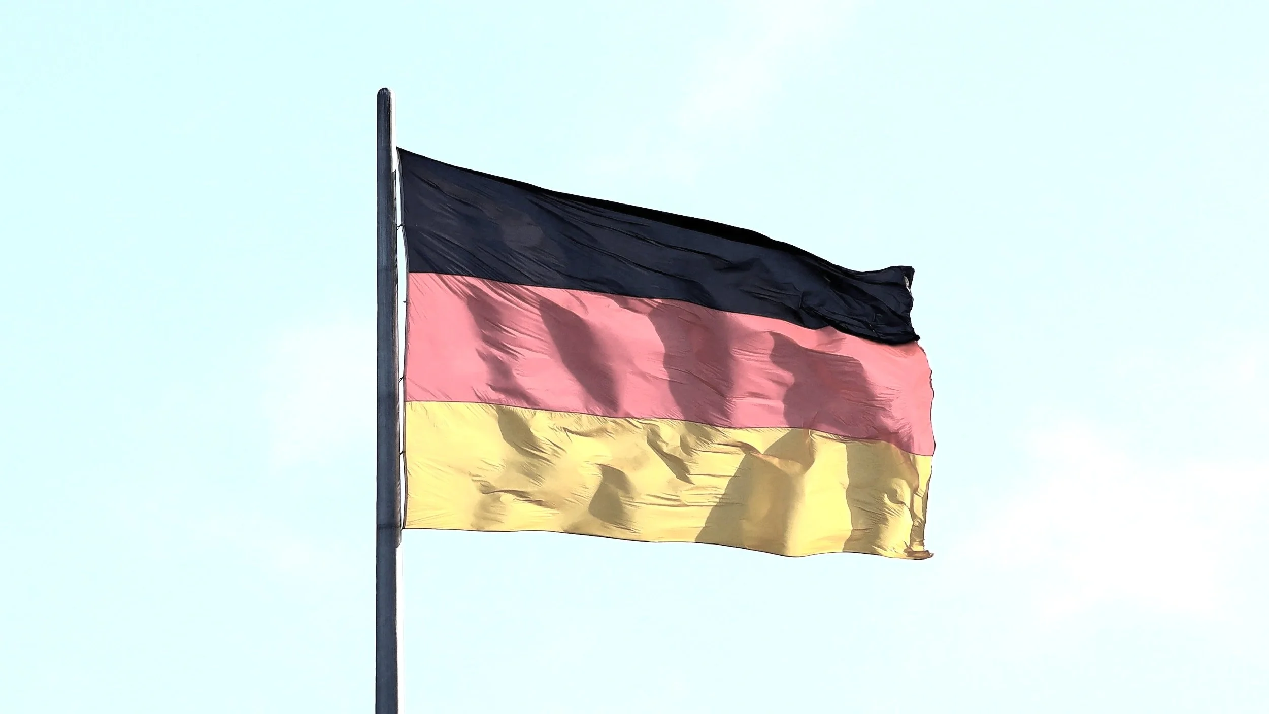 Bandera de Alemania ondeando en el viento, con franjas horizontales de negro, rojo y amarillo, en un mástil contra un cielo despejado.