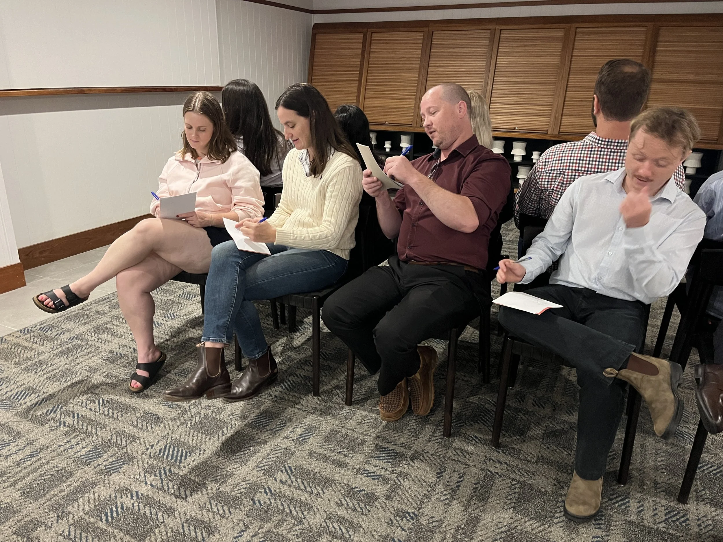 People sitting on chairs in a meeting room, reading or writing notes, with some engaging in conversation, in a professional setting.