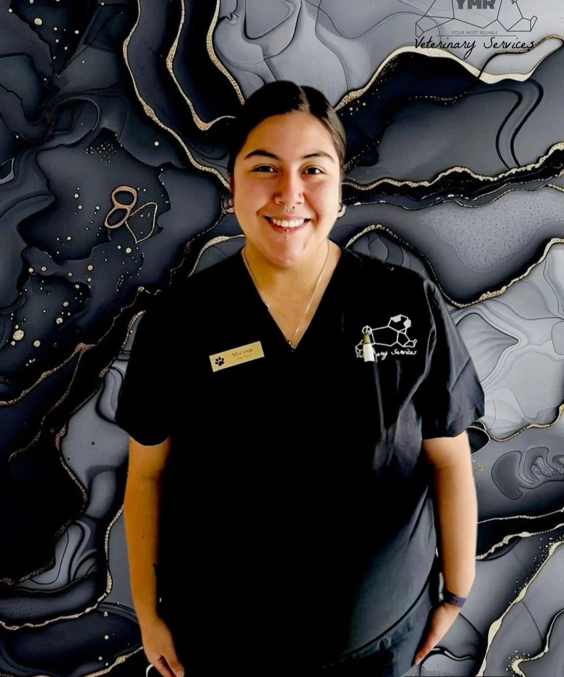 A smiling woman wearing a black uniform with a gold name tag and a veterinary services logo, standing in front of an abstract black and gray wall with gold accents.