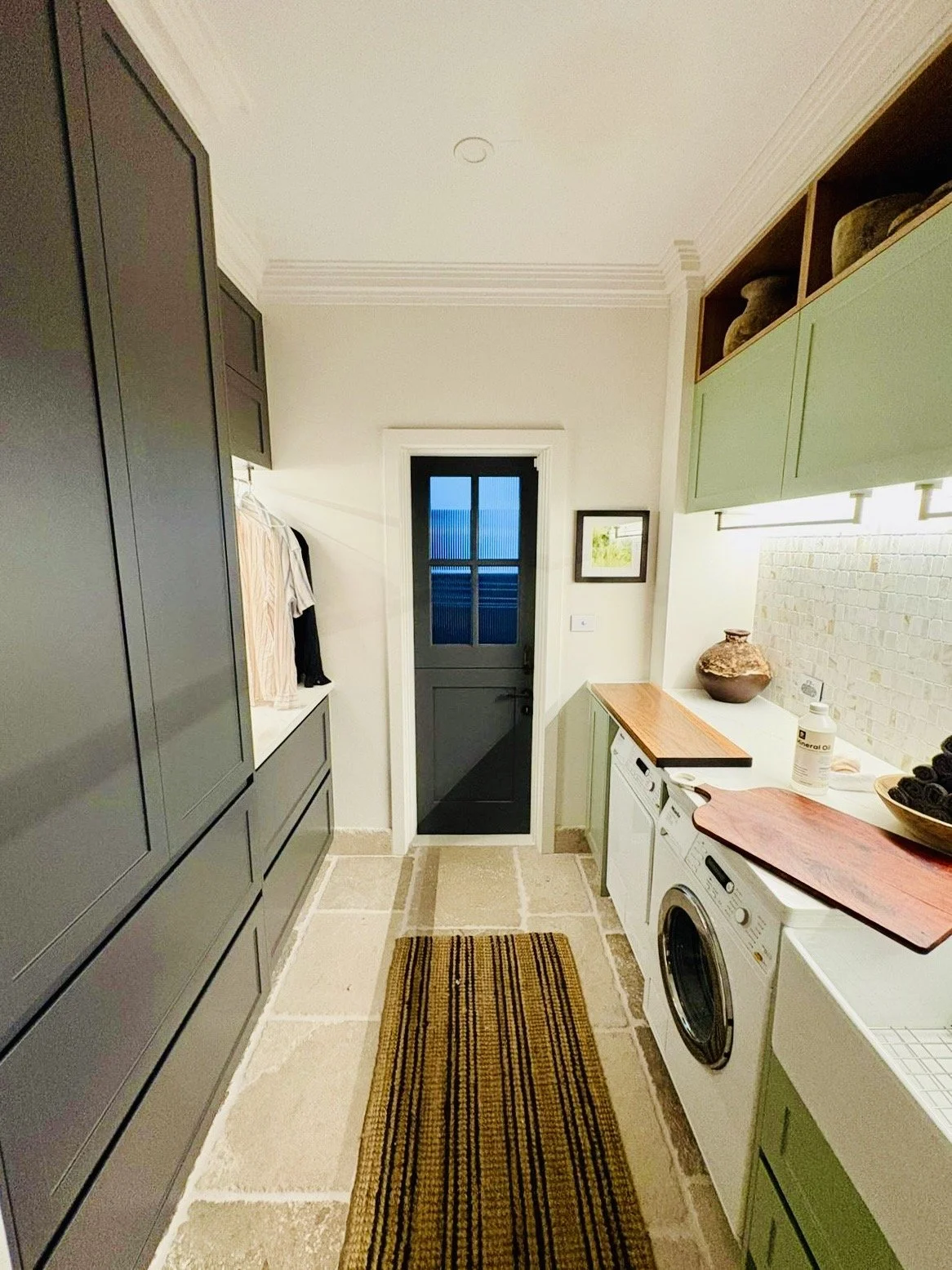 A laundry room with gray cabinetry, a front-loading washing machine, green shelves, a wooden countertop, a brown vase, and a door at the back leading outside.