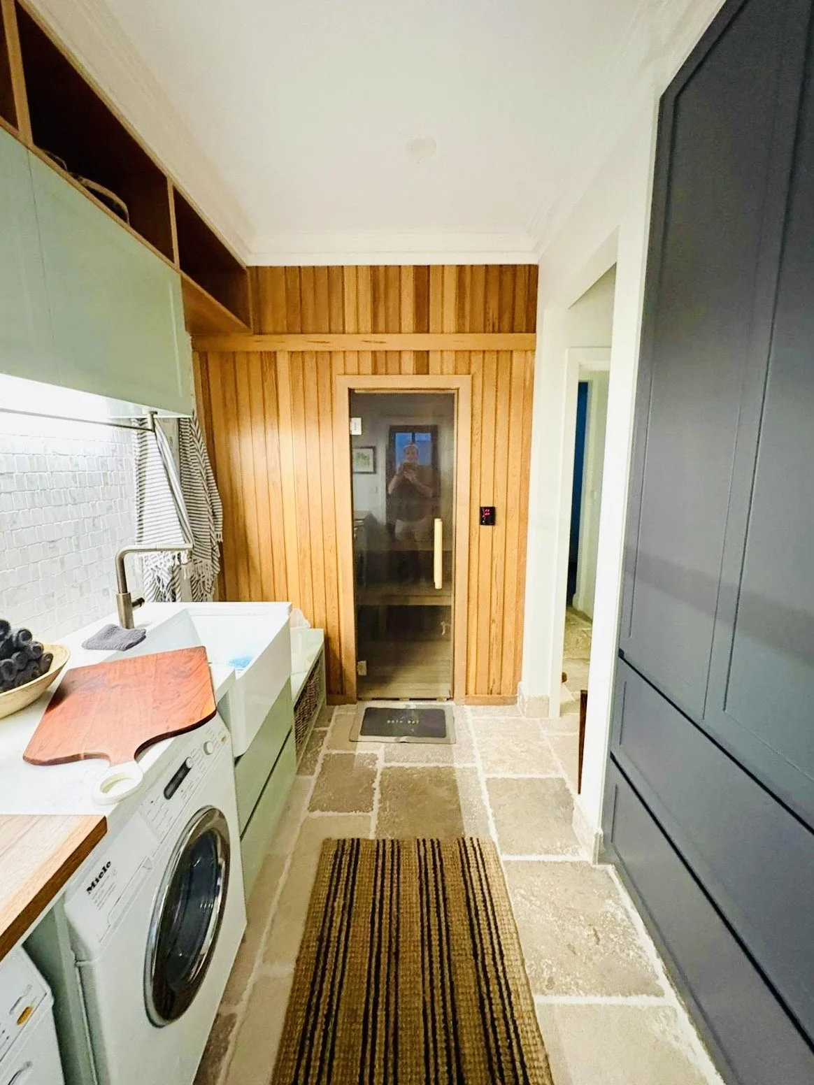 A laundry room with a washing machine on the left, a countertop with a cutting board, a sink, and a bowl of fruit. The room has a stone floor, wooden wall paneling, and cabinets. At the end, there is a door with a glass panel leading to a sauna or another room, and a person is visible taking the photo in the reflection.