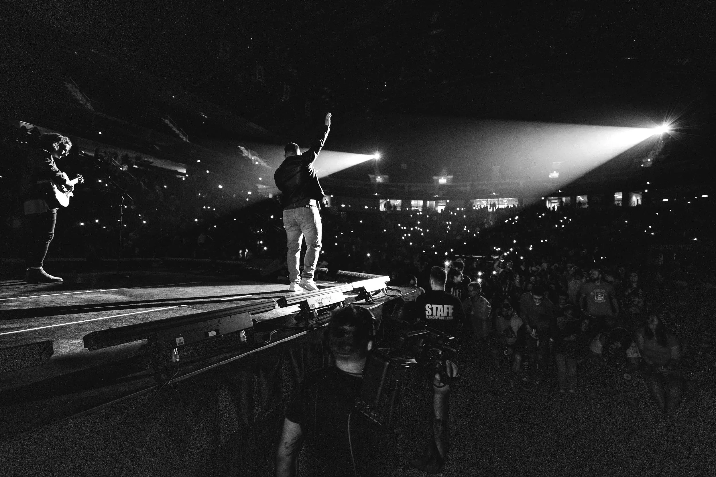 A person on stage waving to the audience with fans and photographers in the crowd, performing under bright stage lights in a large indoor concert venue.