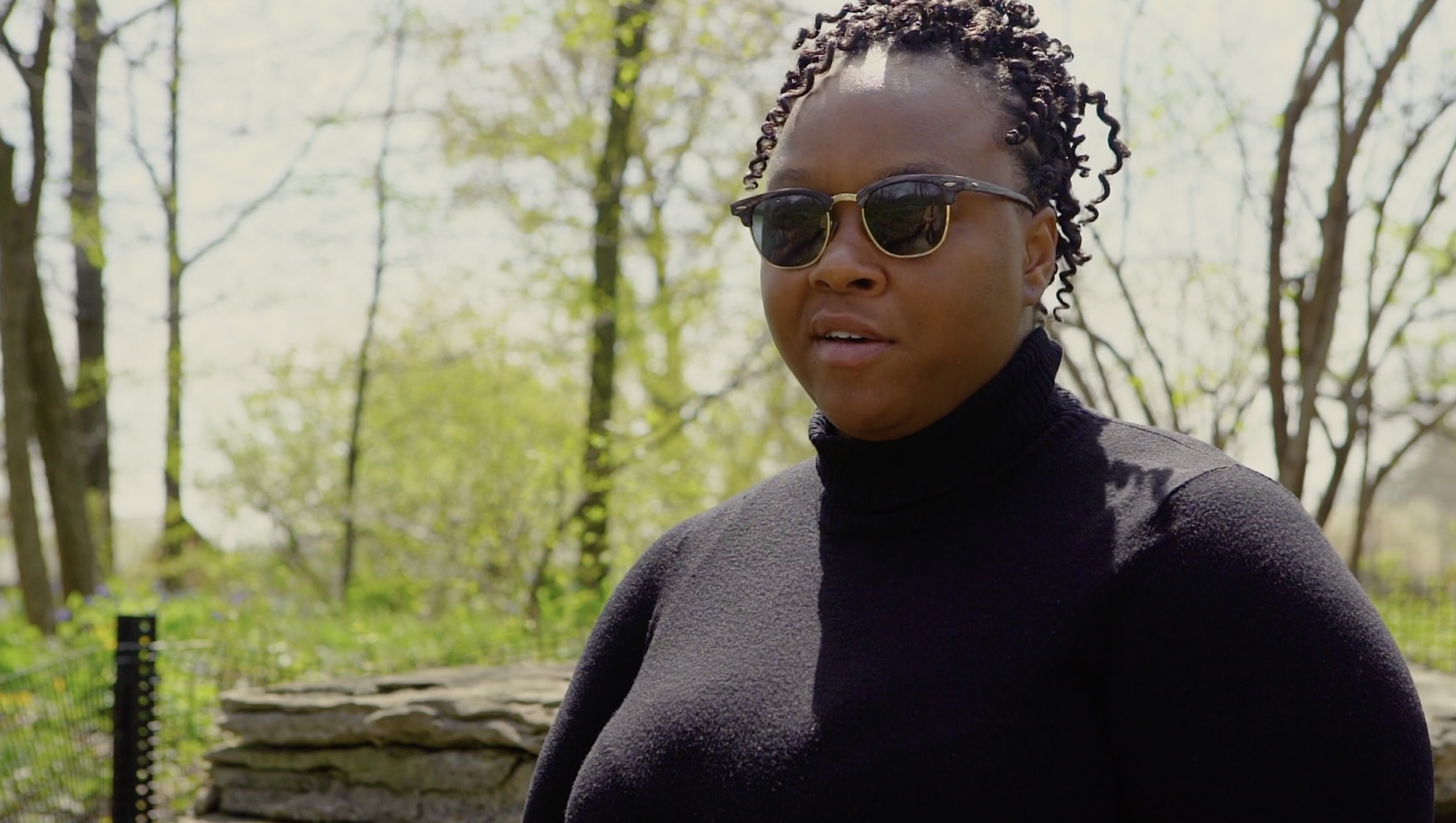 A woman wearing black sunglasses and a black turtleneck sweater outdoors surrounded by trees with green leaves.