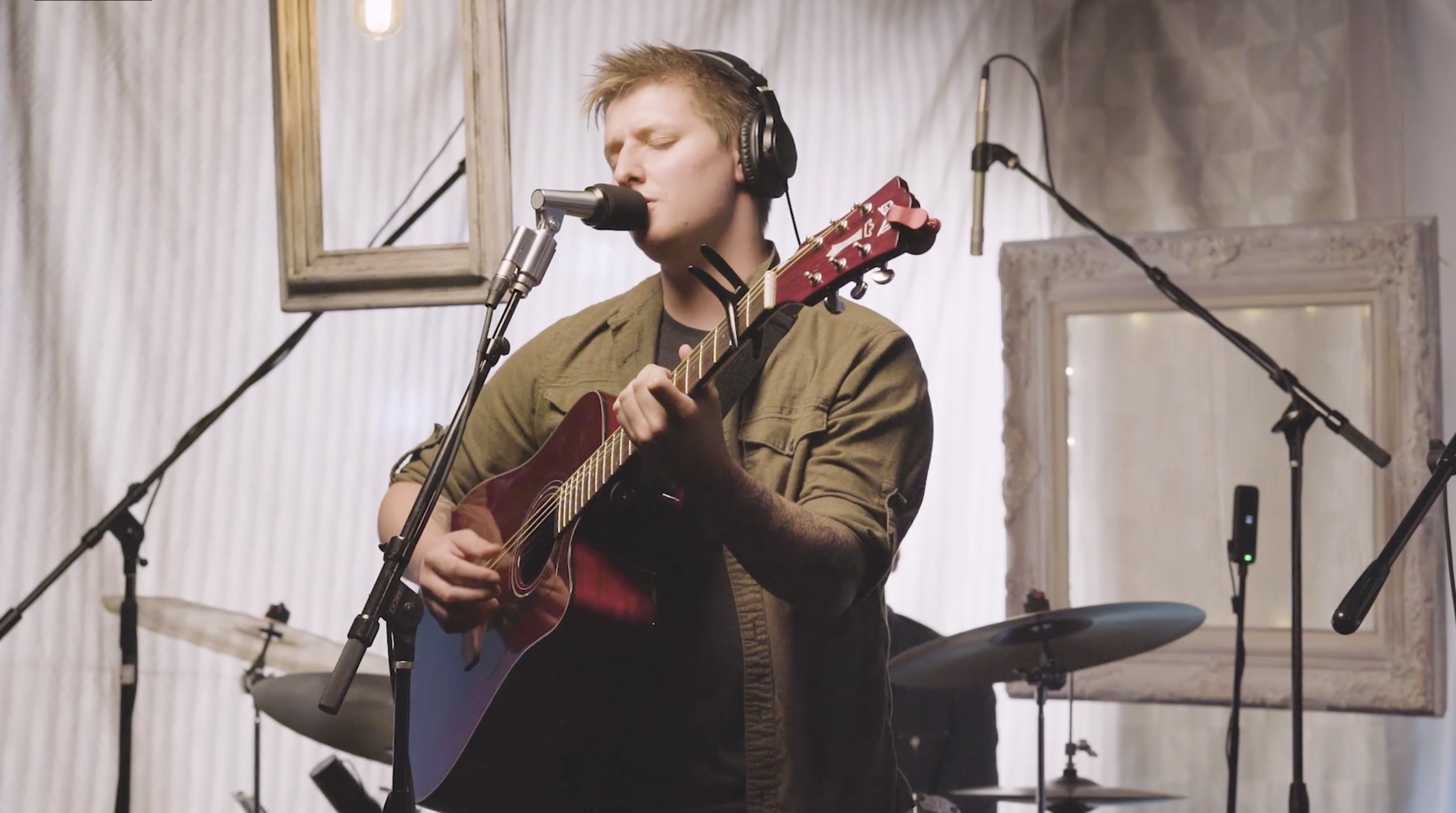A young man with headphones playing an acoustic guitar and singing in a recording studio.