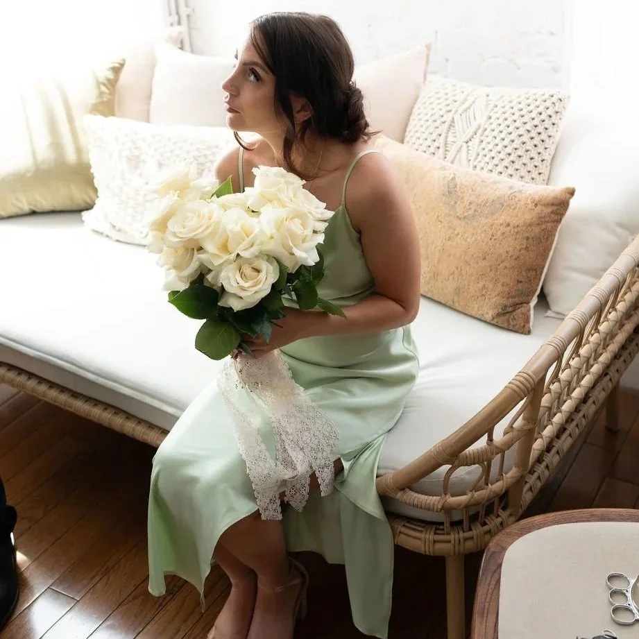 Woman holding a bouquet of white roses, sitting on a wicker bench in a bright room with white walls and cushions.