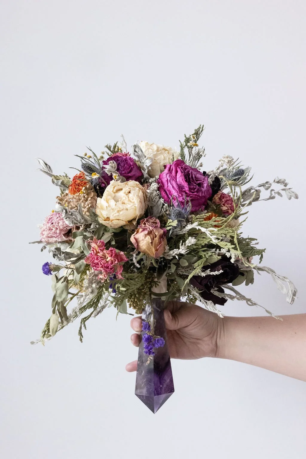 A hand holding a bouquet of dried flowers with various colors including purple, white, pink, and orange, arranged in a purple geometric-shaped vase.
