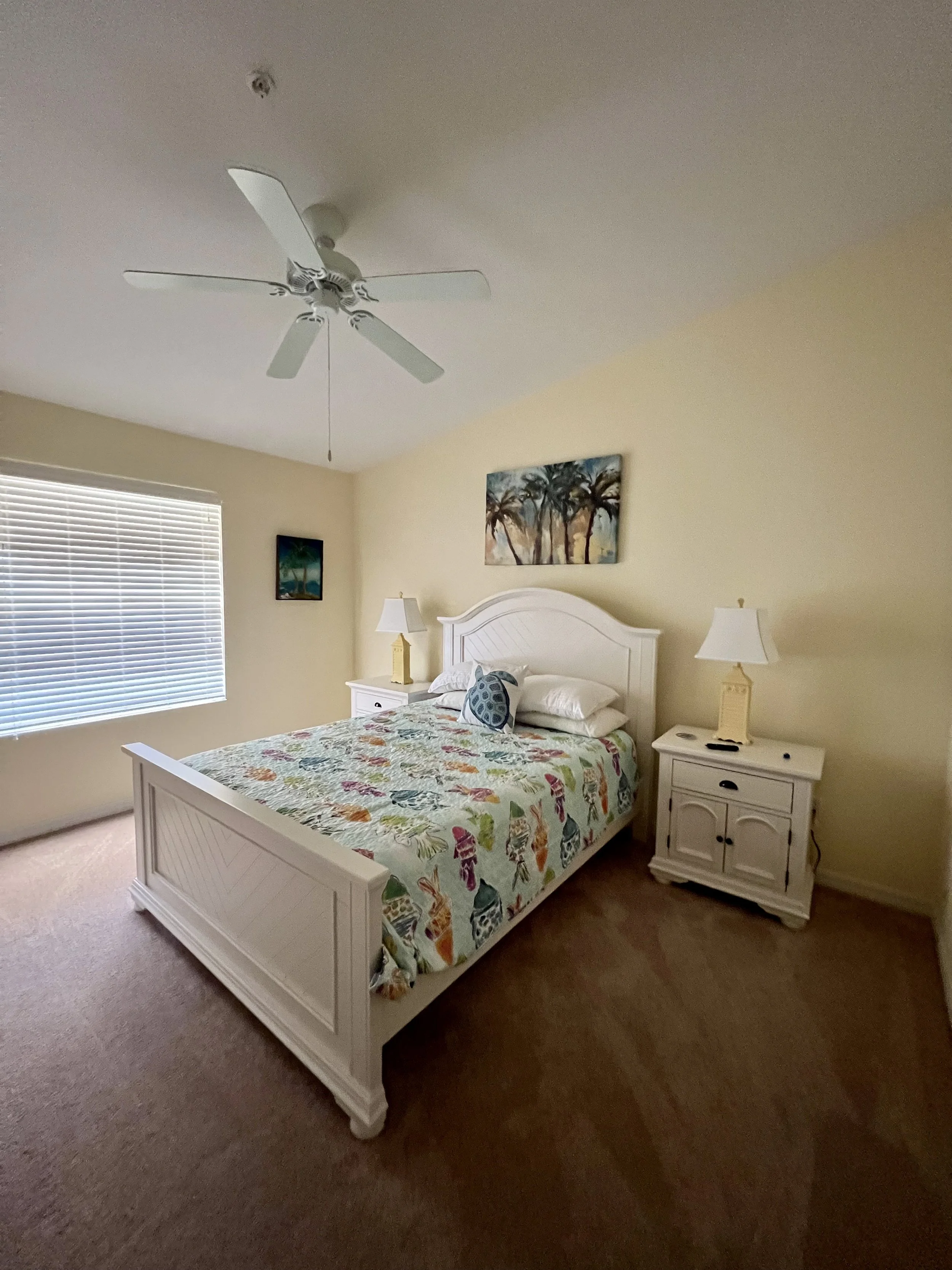 Bedroom with white bed frame, floral bedspread, two nightstands with lamps, paintings on yellow walls, window with blinds, ceiling fan.