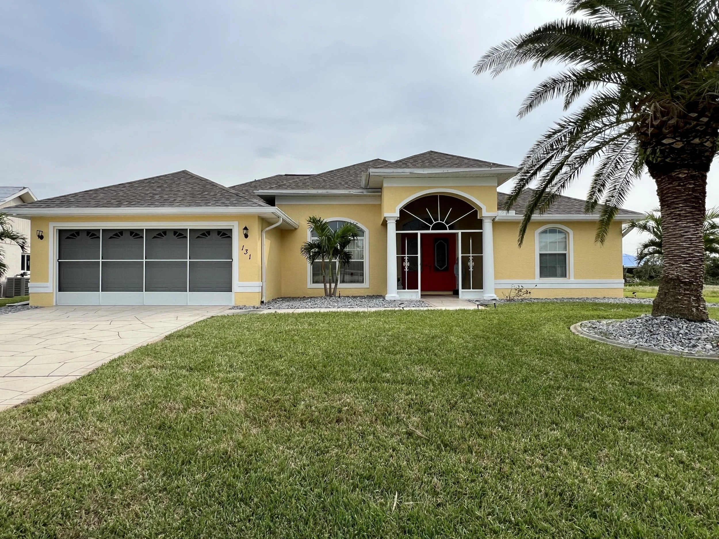 A yellow house with a two-car garage, a red front door, and palm trees in the front yard.