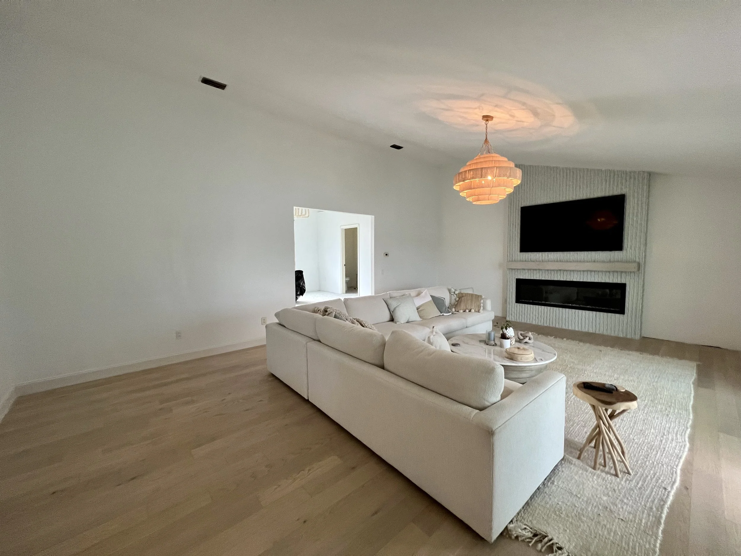 Living room with a white sectional sofa, a coffee table, a rug, a wall-mounted TV above a fireplace, and a pink chandelier hanging from the ceiling.
