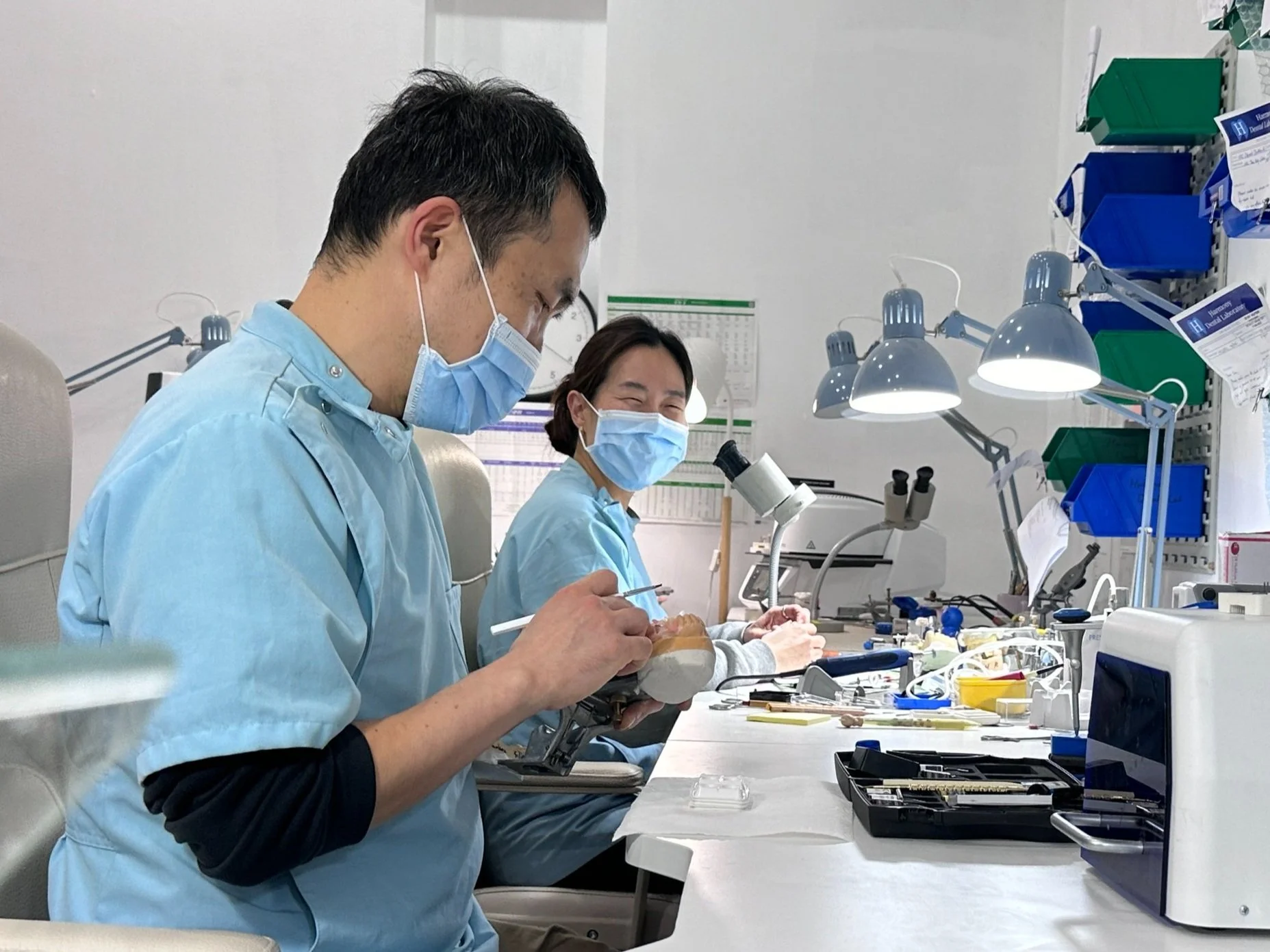 Two dental professionals wearing masks working on a patient in a dental clinic. One professional is using a dental tool, and various dental instruments and equipment are on the worktable.