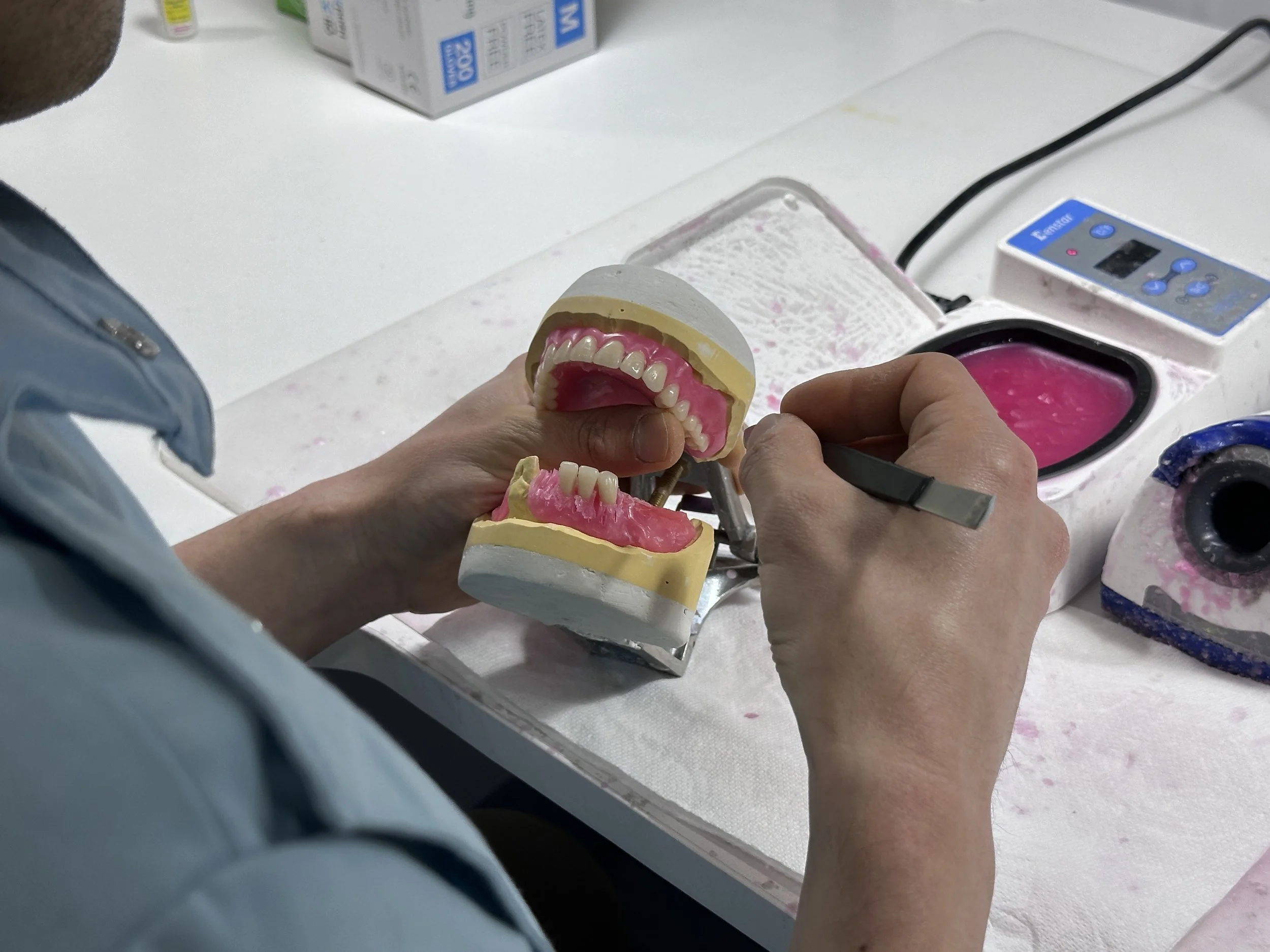 A person working on a dental model with artificial teeth, using a tool at a workstation with dental equipment and supplies.