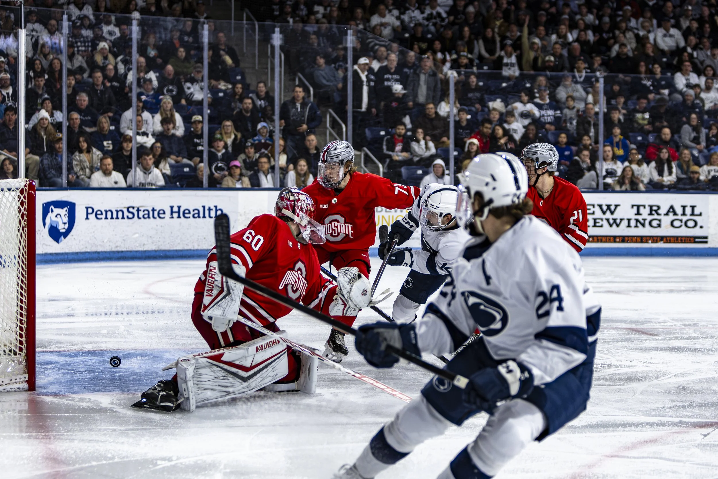Hockey players in action during a game, with the goalie attempting to block a shot and players from both teams skating around the goal area on the ice rink.
