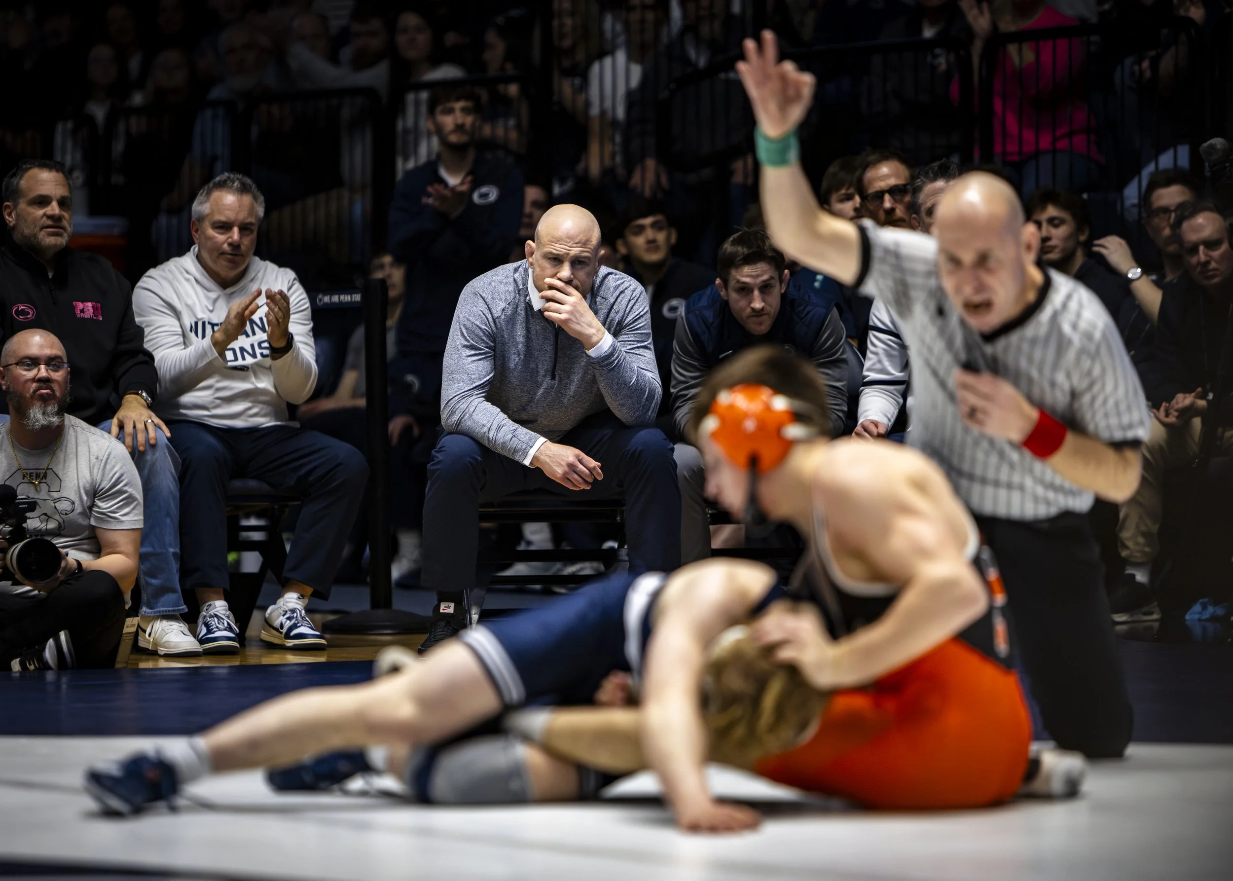 A wrestling match with two competitors on the mat and a referee holding one athlete down, while spectators watch, with some looking concerned and others clapping.