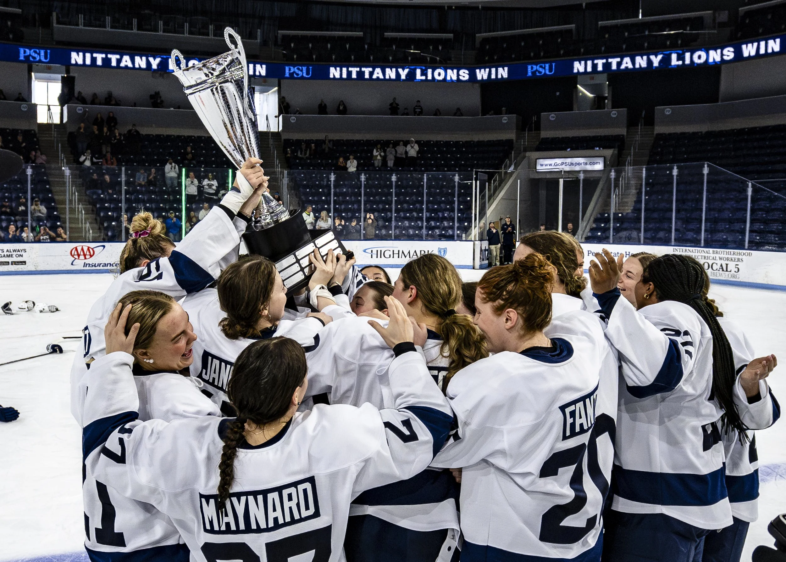 Women's ice hockey team celebrating victory by lifting trophy in an ice rink, with scoreboard and audience in the background.
