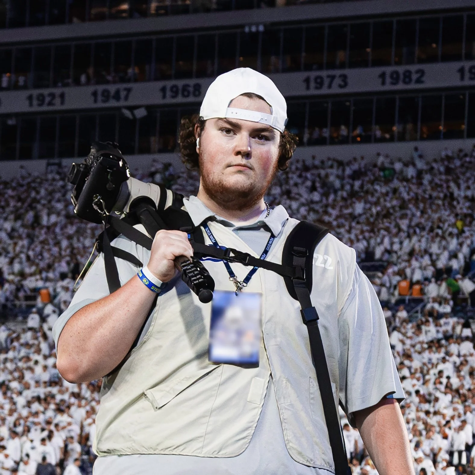 A young male photographer holding a camera on his shoulder at a stadium filled with spectators.