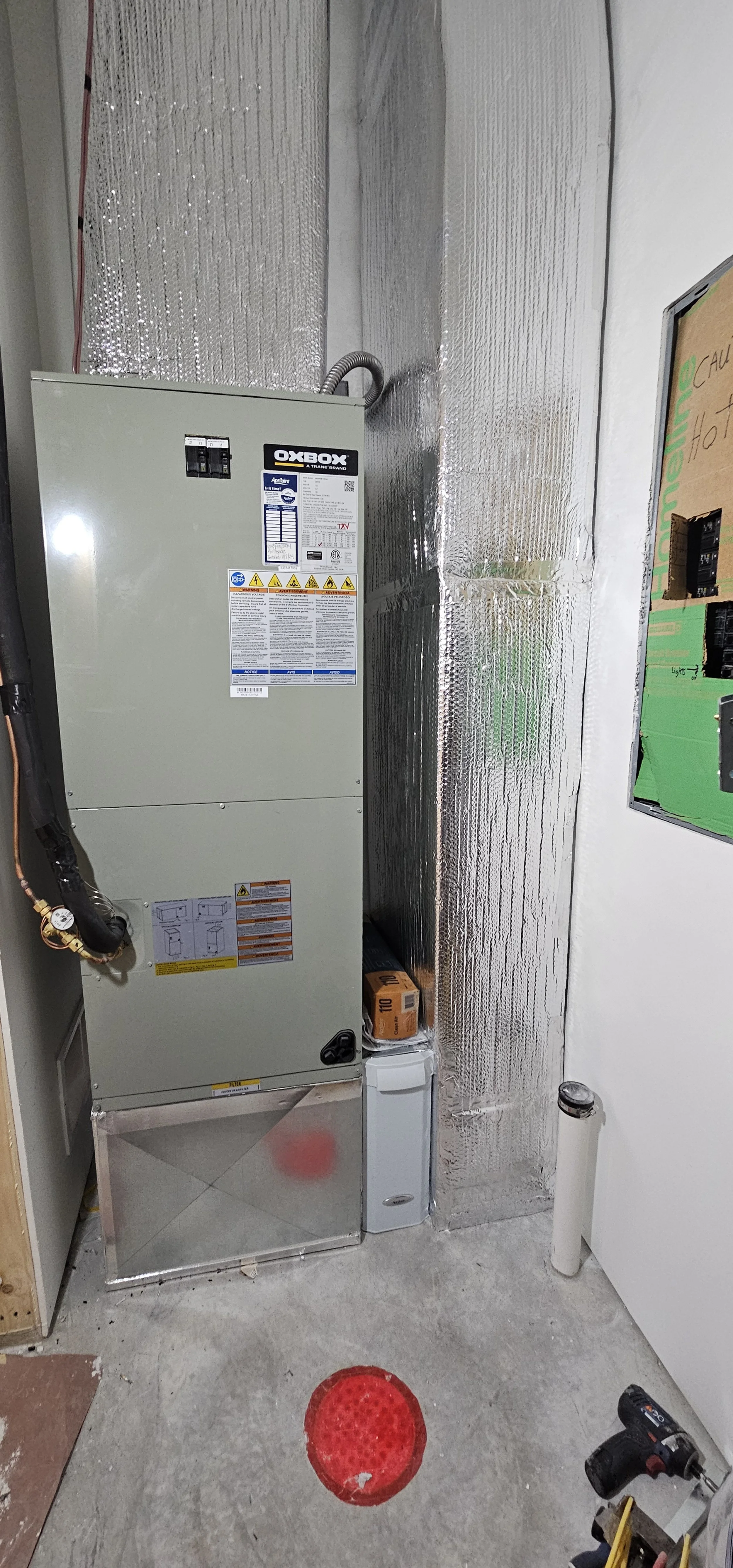 View of an HVAC furnace unit with duct insulation in a utility room, with a red safety cone on the floor, and a drill and construction tools nearby.