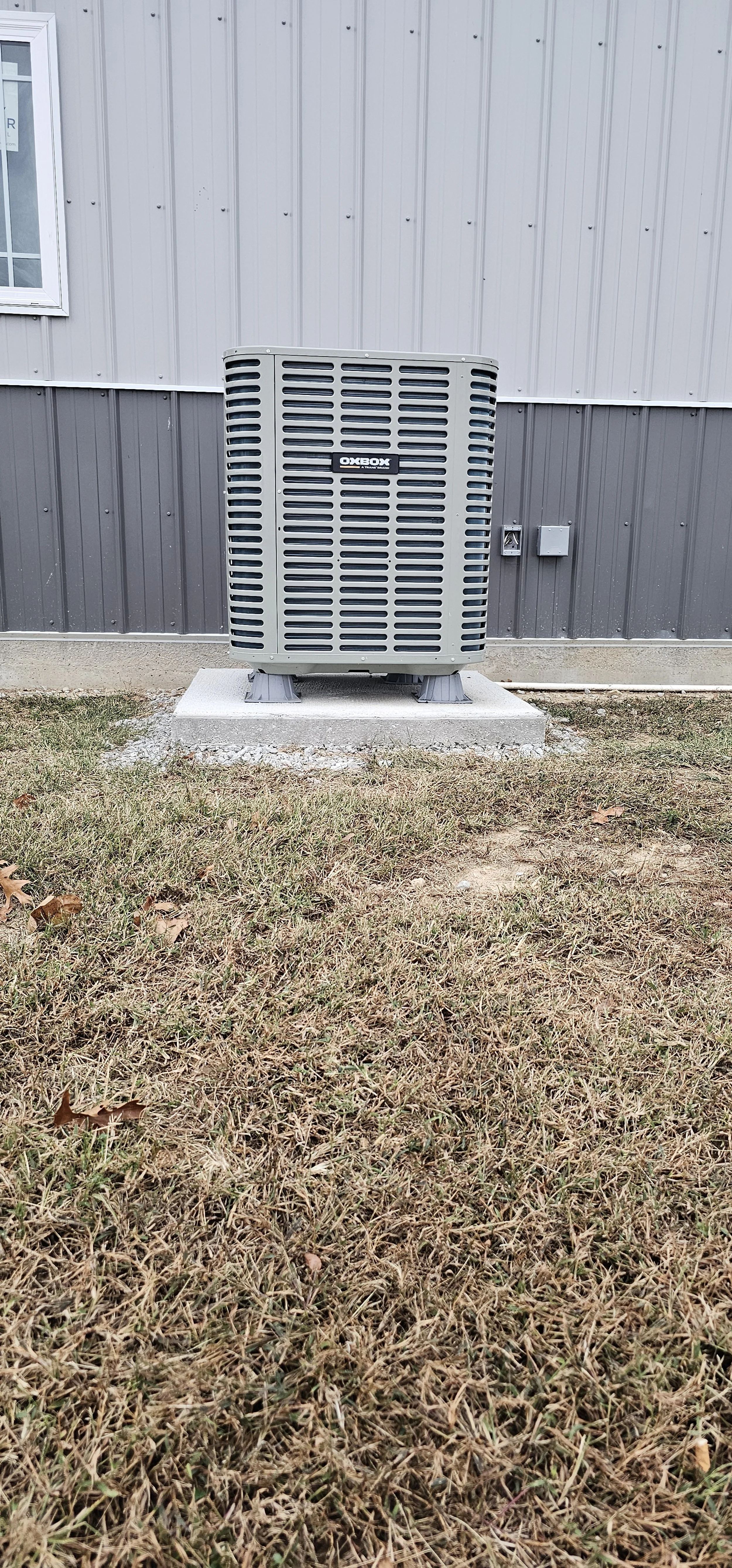 An outdoor air conditioning unit on a concrete pad next to a metal-sided building with a window and electrical fixtures, on a patchy grassy area.