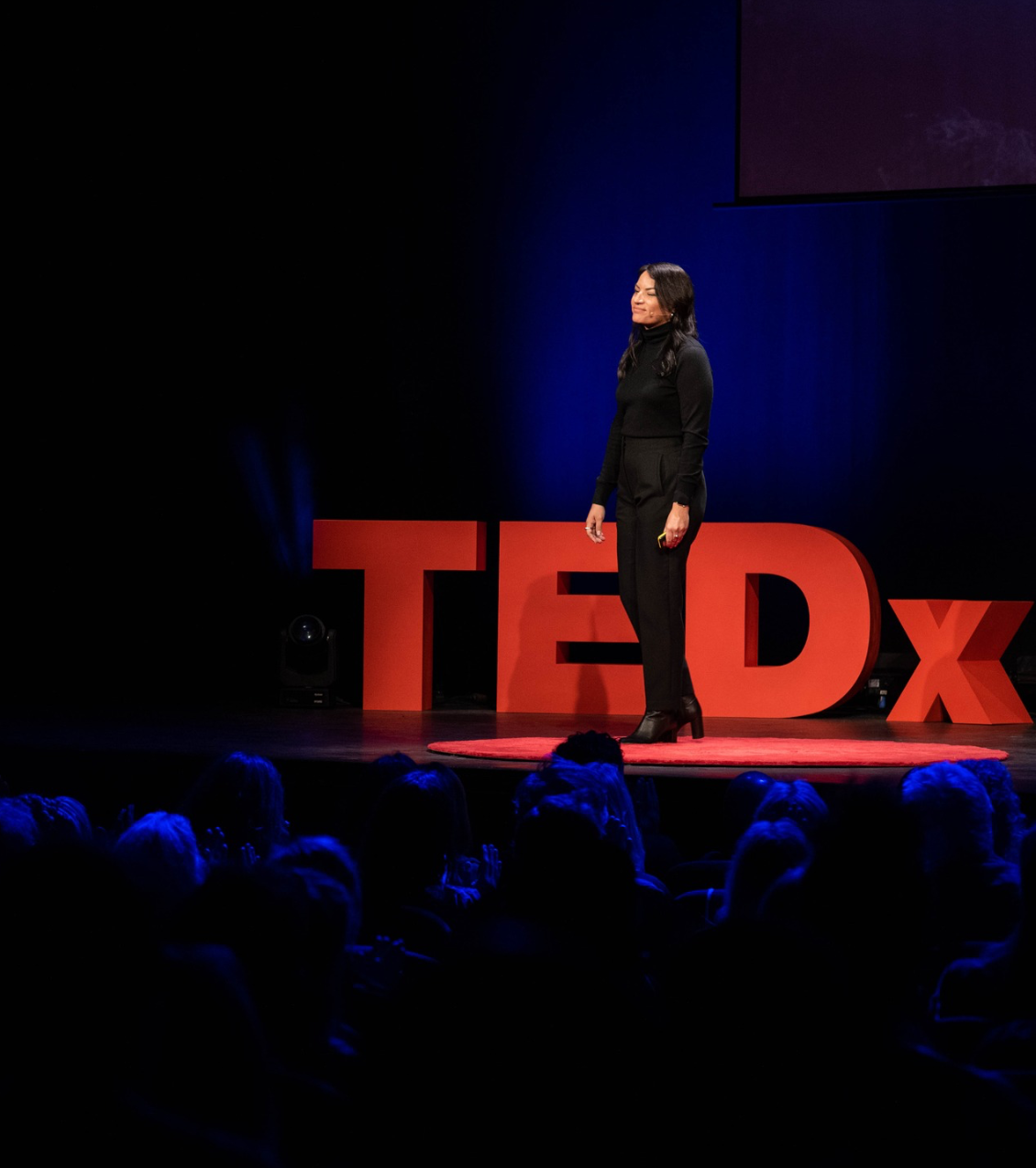 Neliana Fuenmayor, CEO of OPEN.MODE, stands on a dark stage delivering a TEDx talk. Large red "TEDx" letters are behind her. She wears a black turtleneck and trousers, looking to her right while addressing the audience.