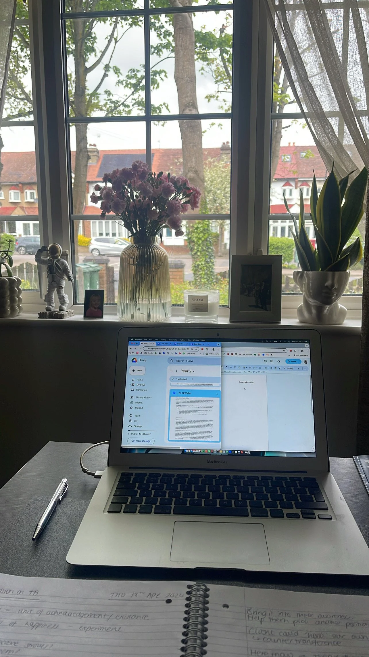 A workspace with a MacBook Air laptop on a table, an open notebook with handwritten notes, and a pen. Behind the laptop, there's a window with a view of trees and houses outside. The windowsill holds a vase with pink flowers, a framed photo, a small figure, a candle, and a potted plant with long green leaves.