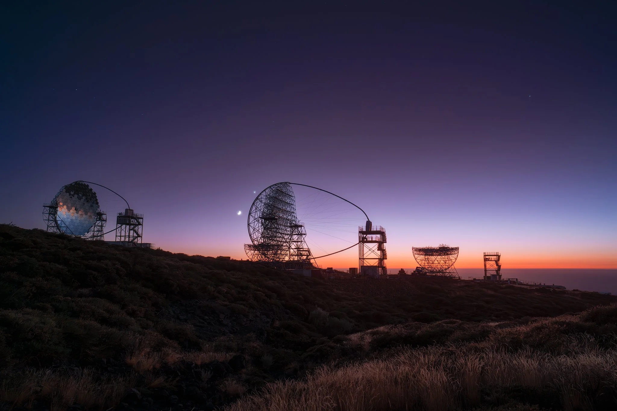 Radio telescopes silhouetted against a sunset sky with stars and planets visible.