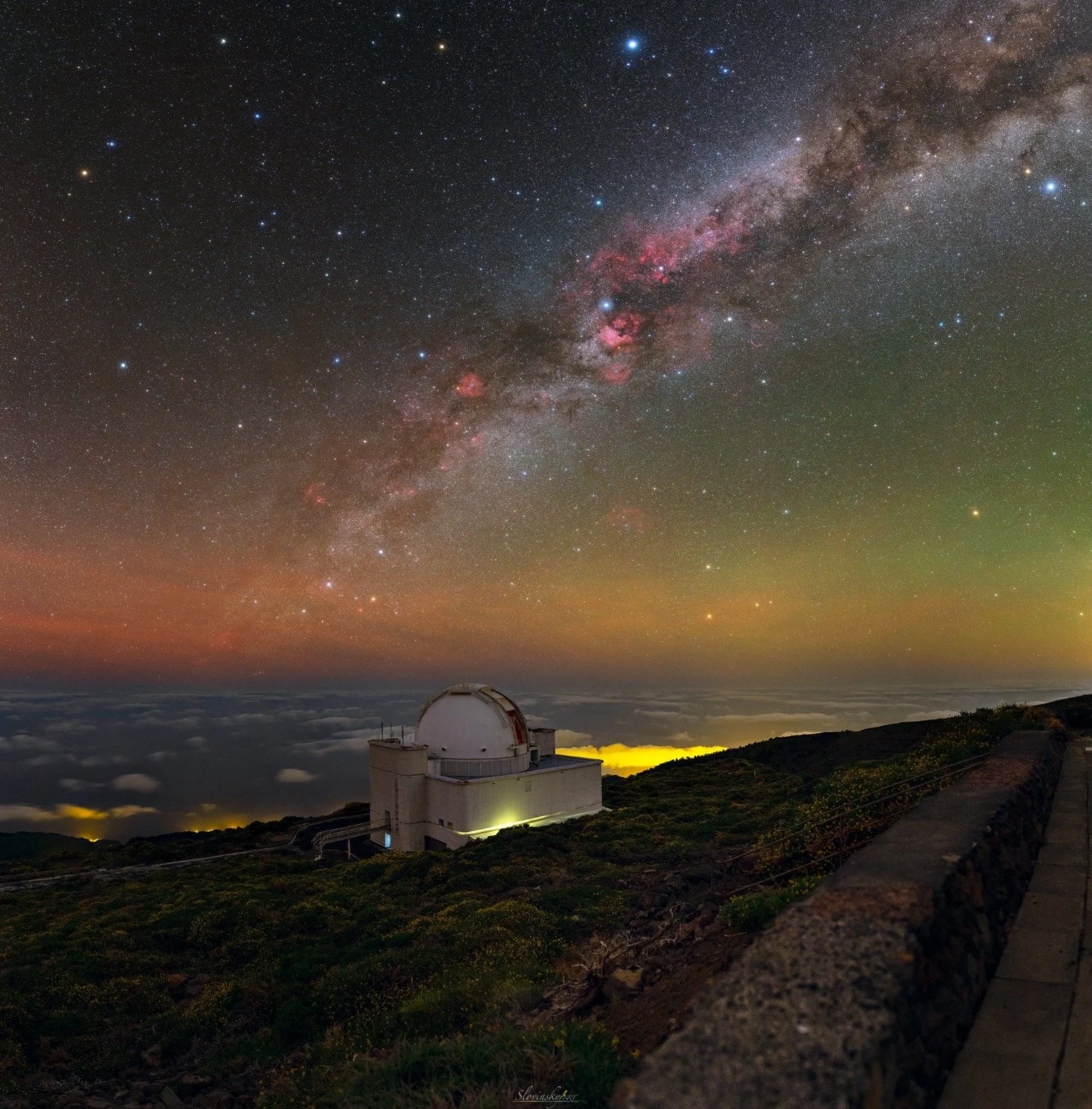 An observatory building on a hillside under a starry night sky with the Milky Way galaxy visible above, and clouds below the horizon.