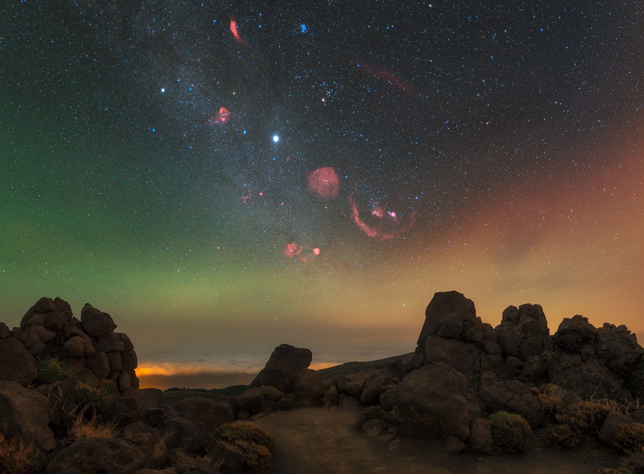 Night sky filled with stars, including the Milky Way galaxy and pink-colored nebula, above rocky landscape with scattered boulders and a distant glow.