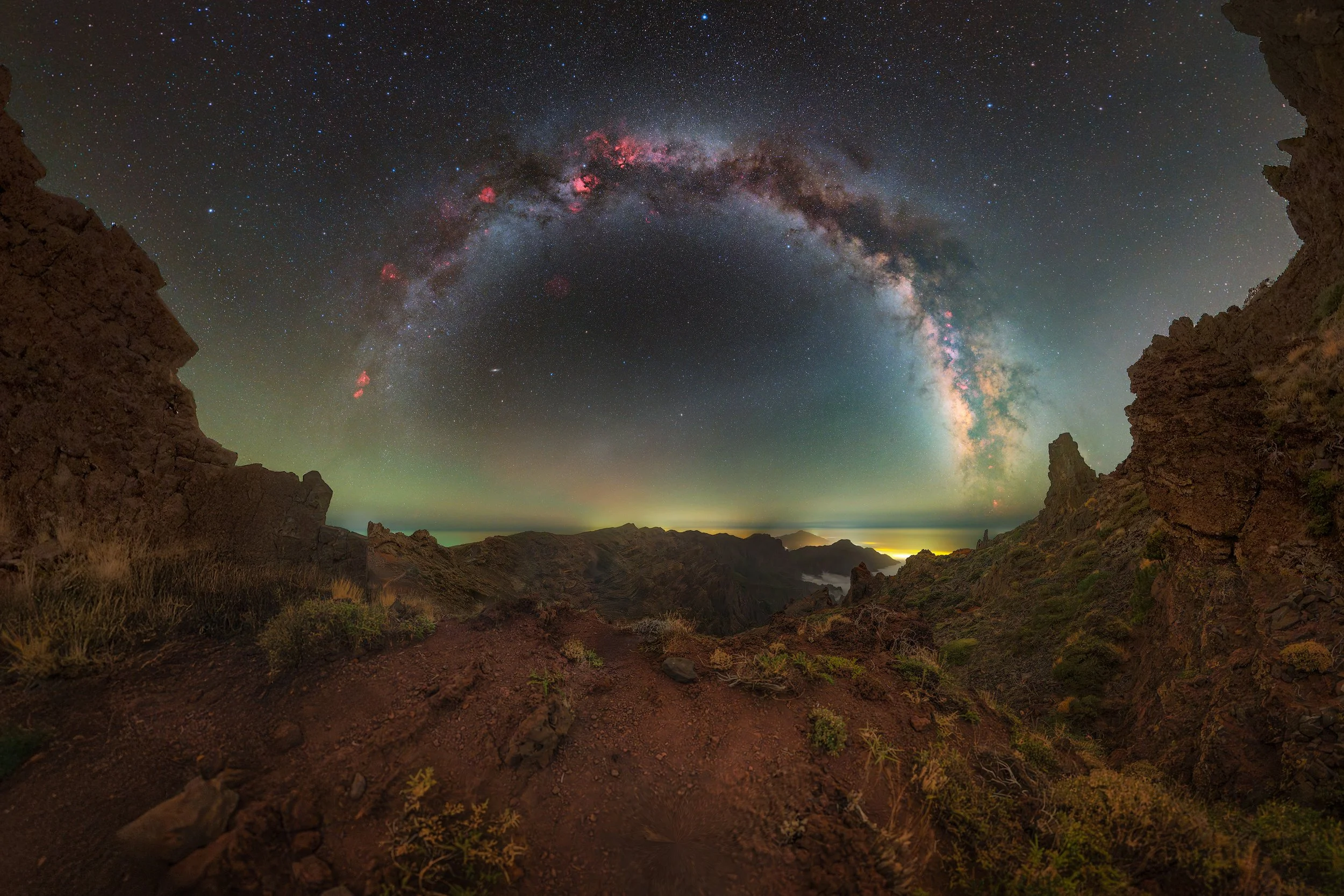 A night sky with the Milky Way galaxy arching over rugged rocky terrain and mountains, illuminated by faint green and yellowish light on the horizon.