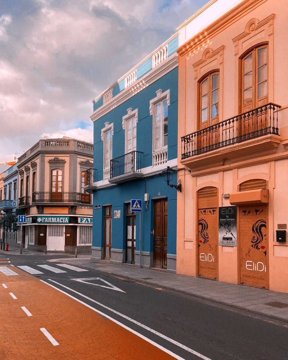 Colorful buildings lining a street with a pharmacy on the corner, pedestrian crosswalk, and a small traffic sign, under a partly cloudy sky
