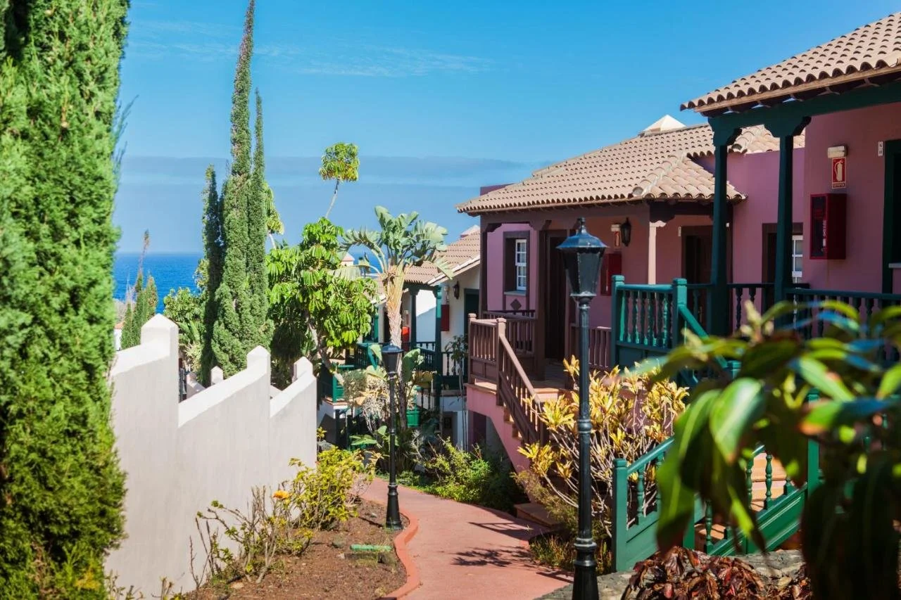 Colorful houses with balconies and stairs in a seaside neighborhood, surrounded by trees and plants, with the ocean visible in the background.