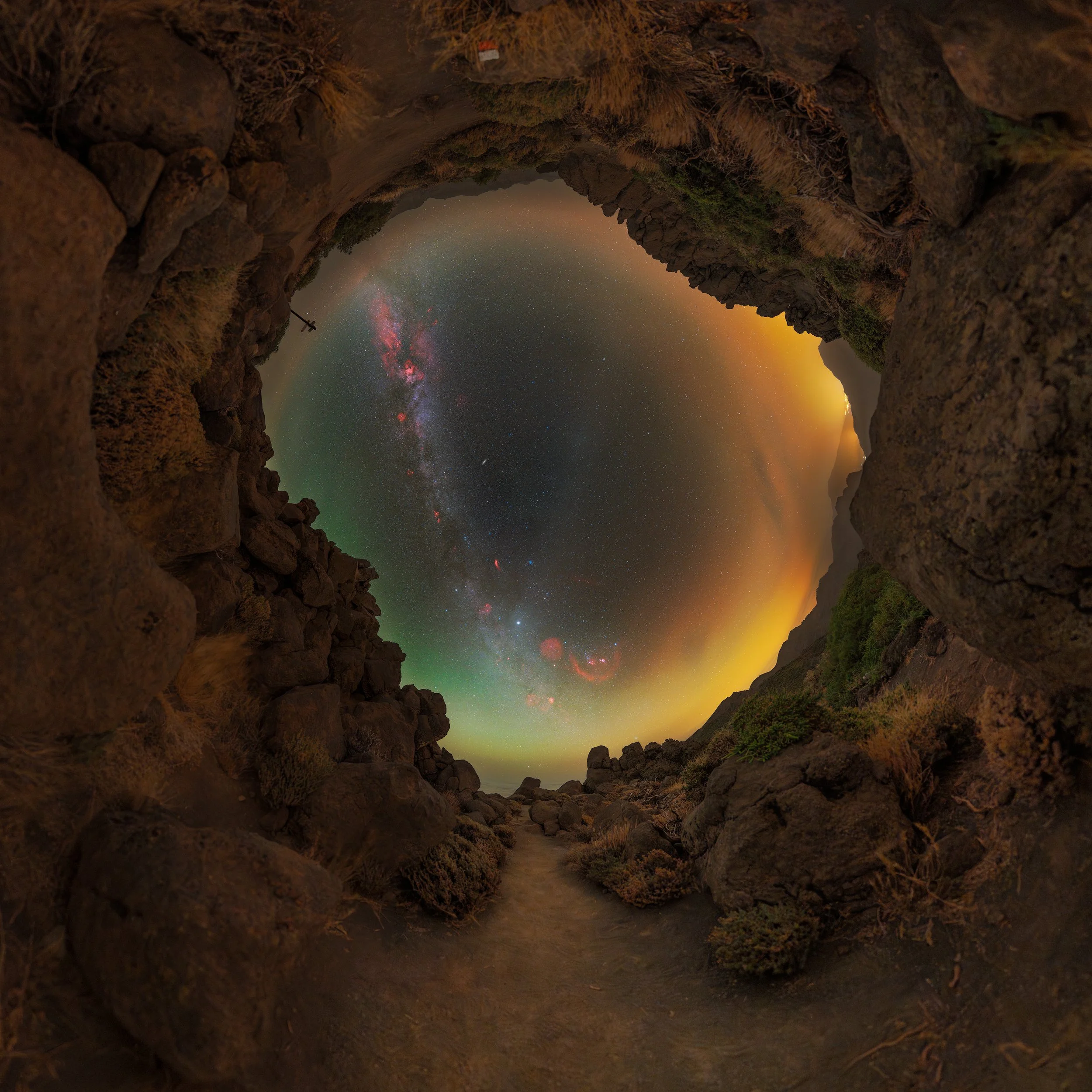 A circular view of the night sky with the Milky Way galaxy, colorful auroras, and stars, framed by rocky terrain and sparse vegetation.