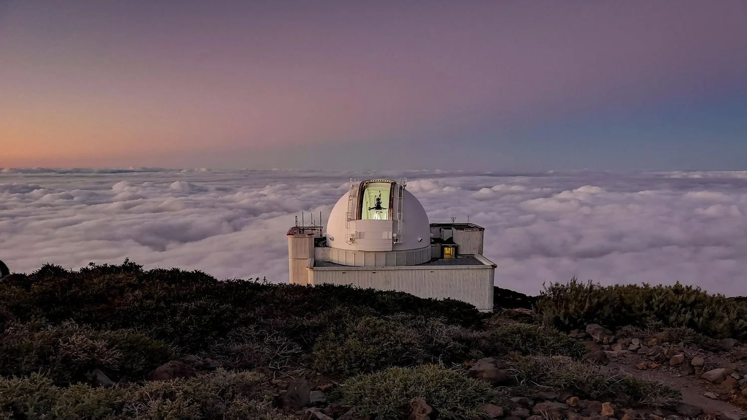 A large astronomical observatory with a domed telescope around sunset, above a sea of clouds and mountain terrain.