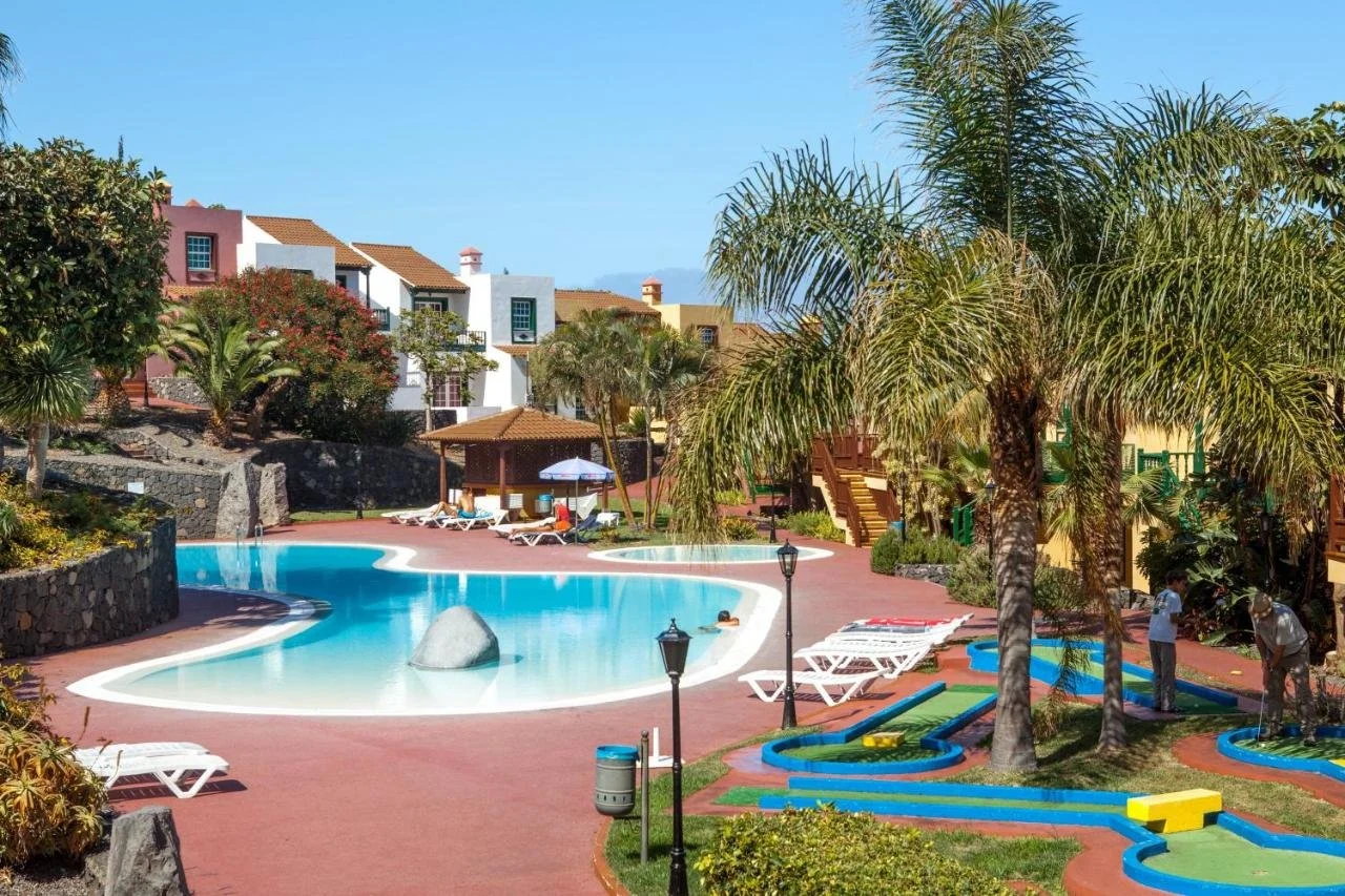 Resort pool area with lounge chairs, umbrellas, palm trees, and colorful buildings in the background under a clear blue sky.