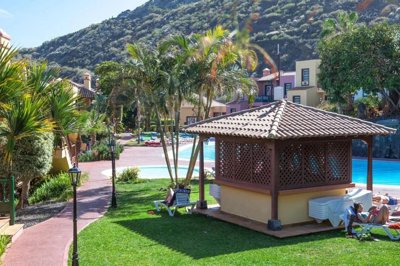 Resort with a swimming pool, palm trees, pathways, and lounge chairs, surrounded by colorful buildings and a mountain in the background.