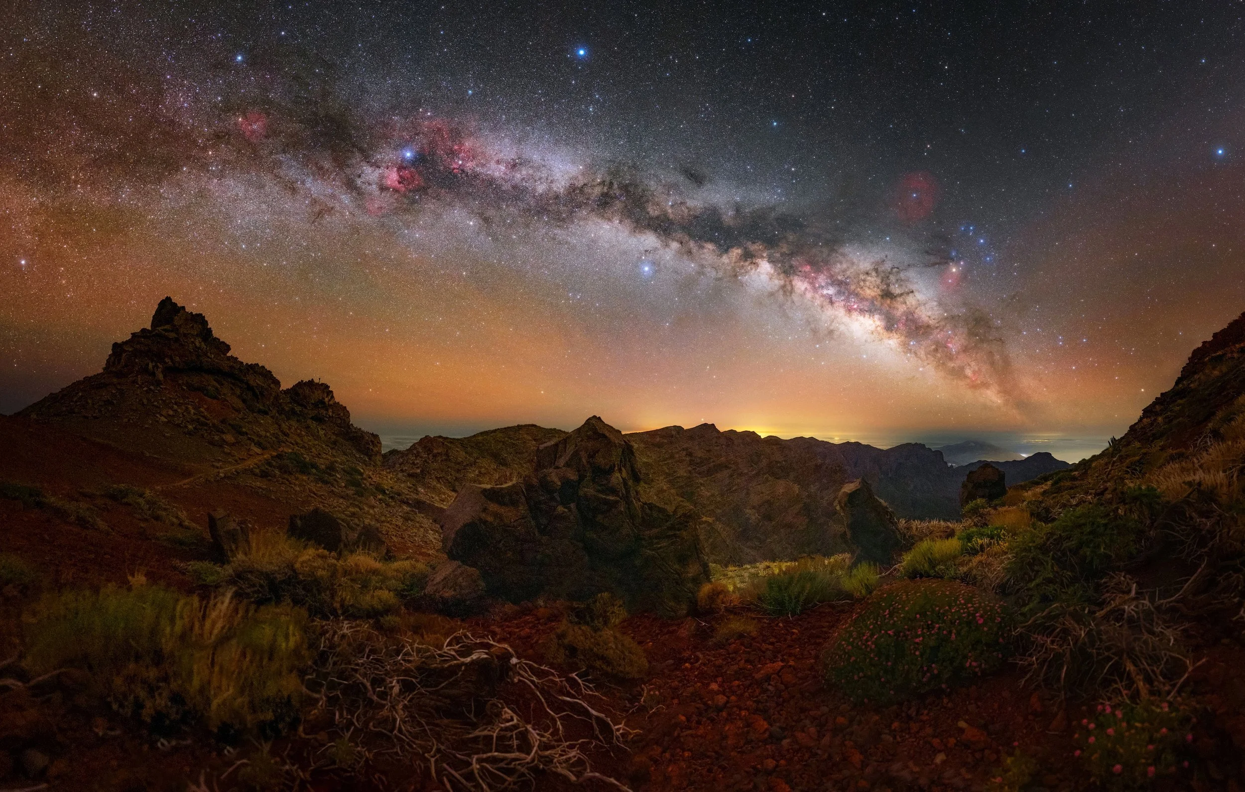 Nighttime view of the Milky Way galaxy above rugged mountains with sparse vegetation and a reddish-orange glow on the horizon.