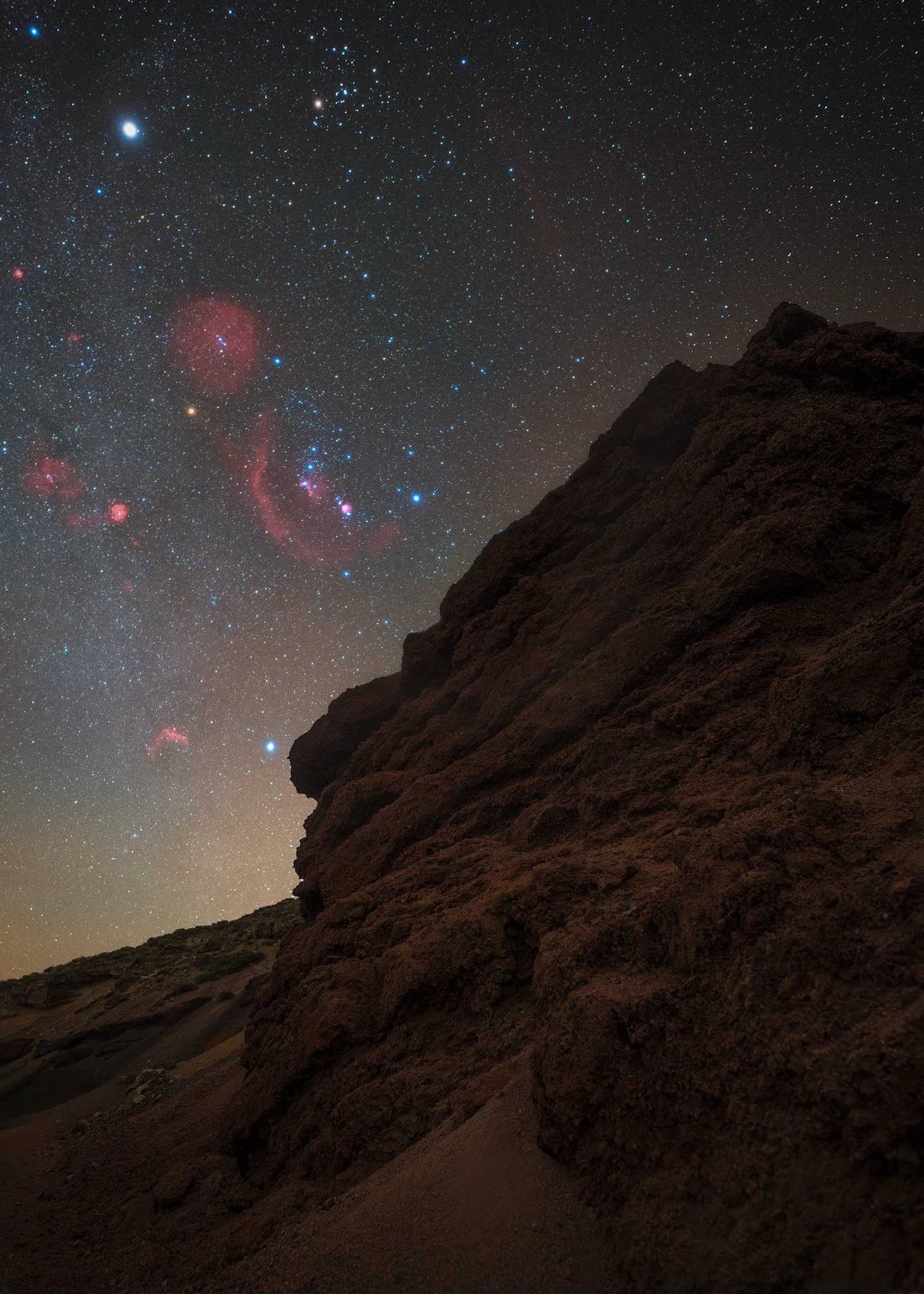 Night sky filled with stars, planets, and nebulae above a dark, rocky mountain slope.