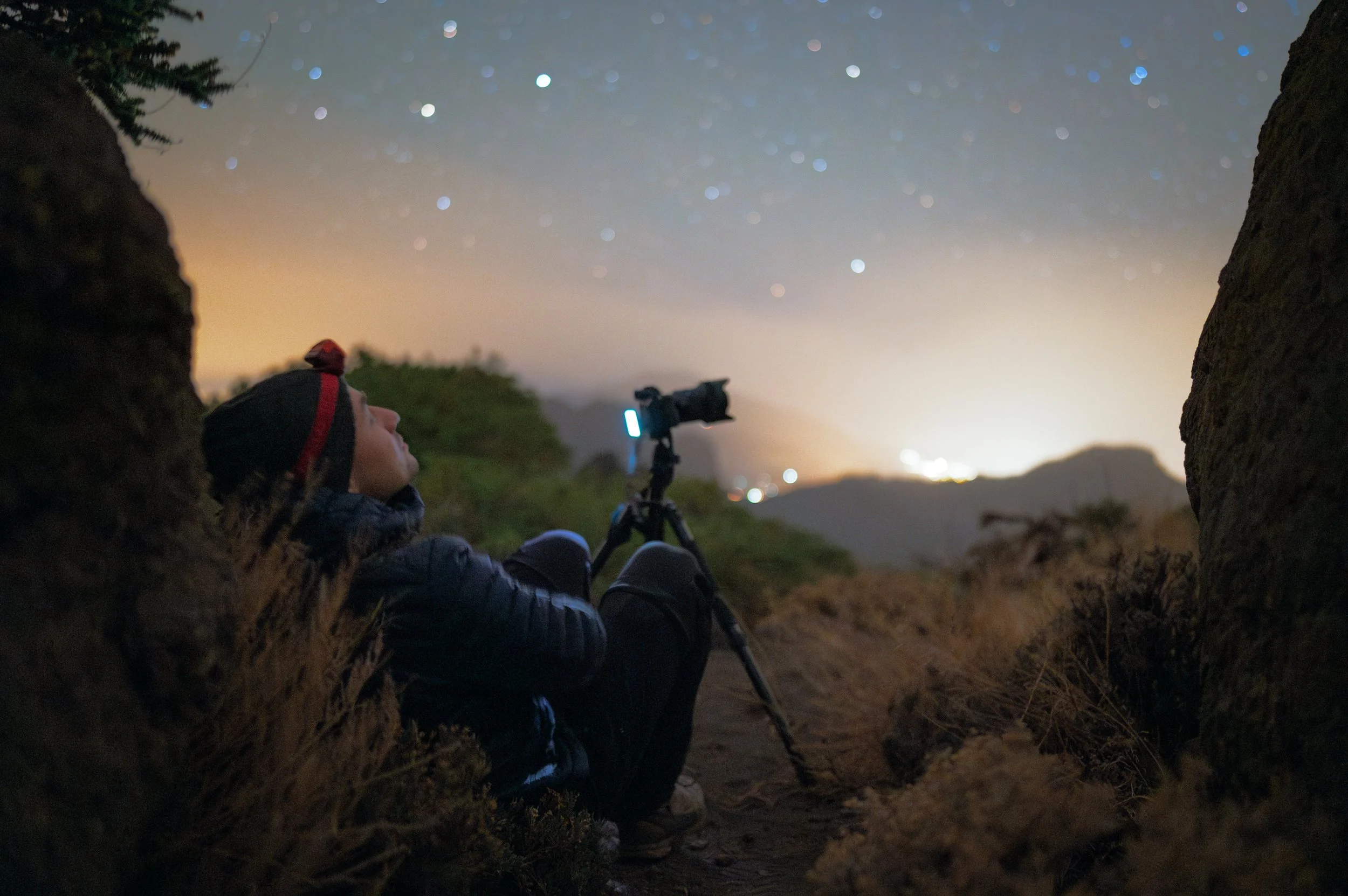 Person sitting on the ground between rocks at night, looking up at the starry sky with a telescope set up nearby.