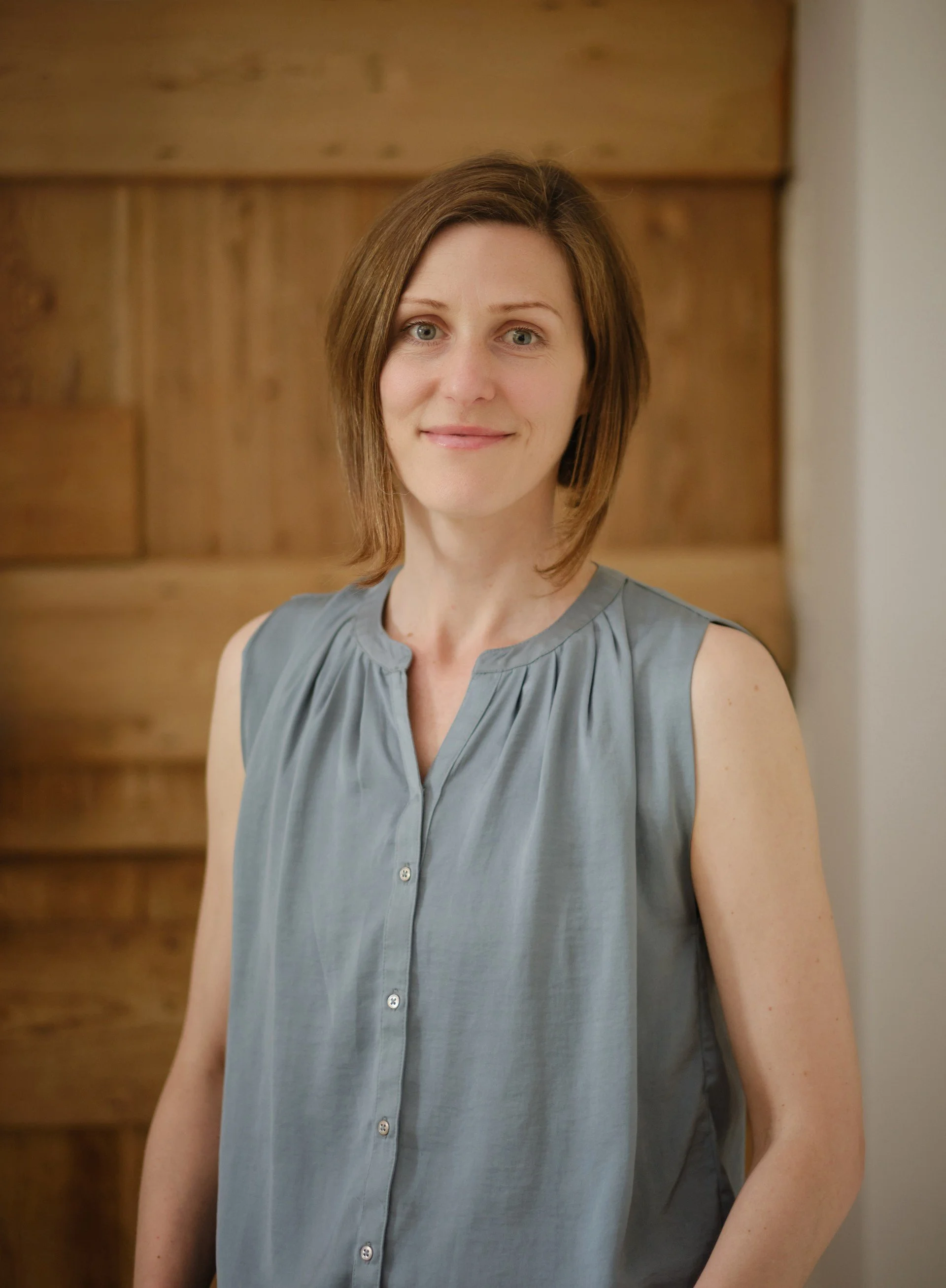 A woman with shoulder-length brown hair and blue eyes smiling while wearing a sleeveless gray blouse, standing in front of a wooden background.