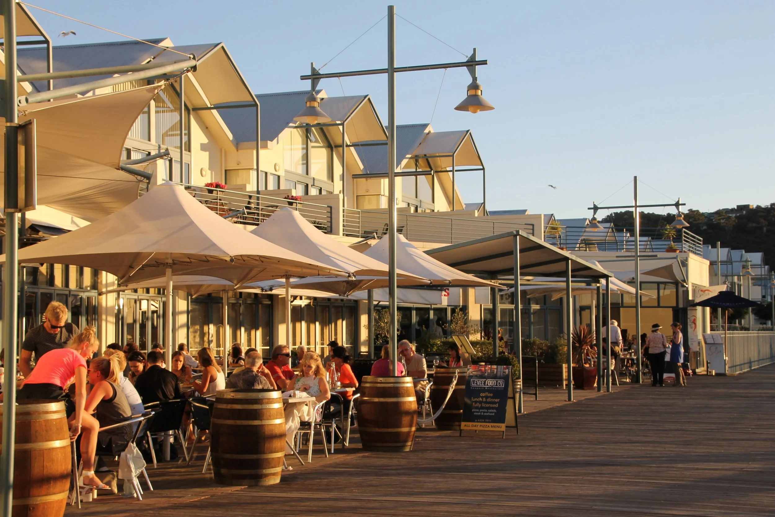 Outdoor restaurant patio filled with people dining under large umbrellas. Overhead are modern light fixtures attached to tall poles. The building behind features large windows and balconies, and there are hills in the background under a clear sky.