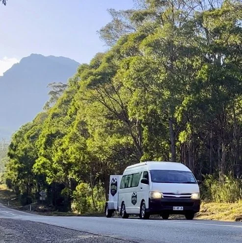 A white police van parked on the side of a rural road surrounded by green trees and mountains in the background.