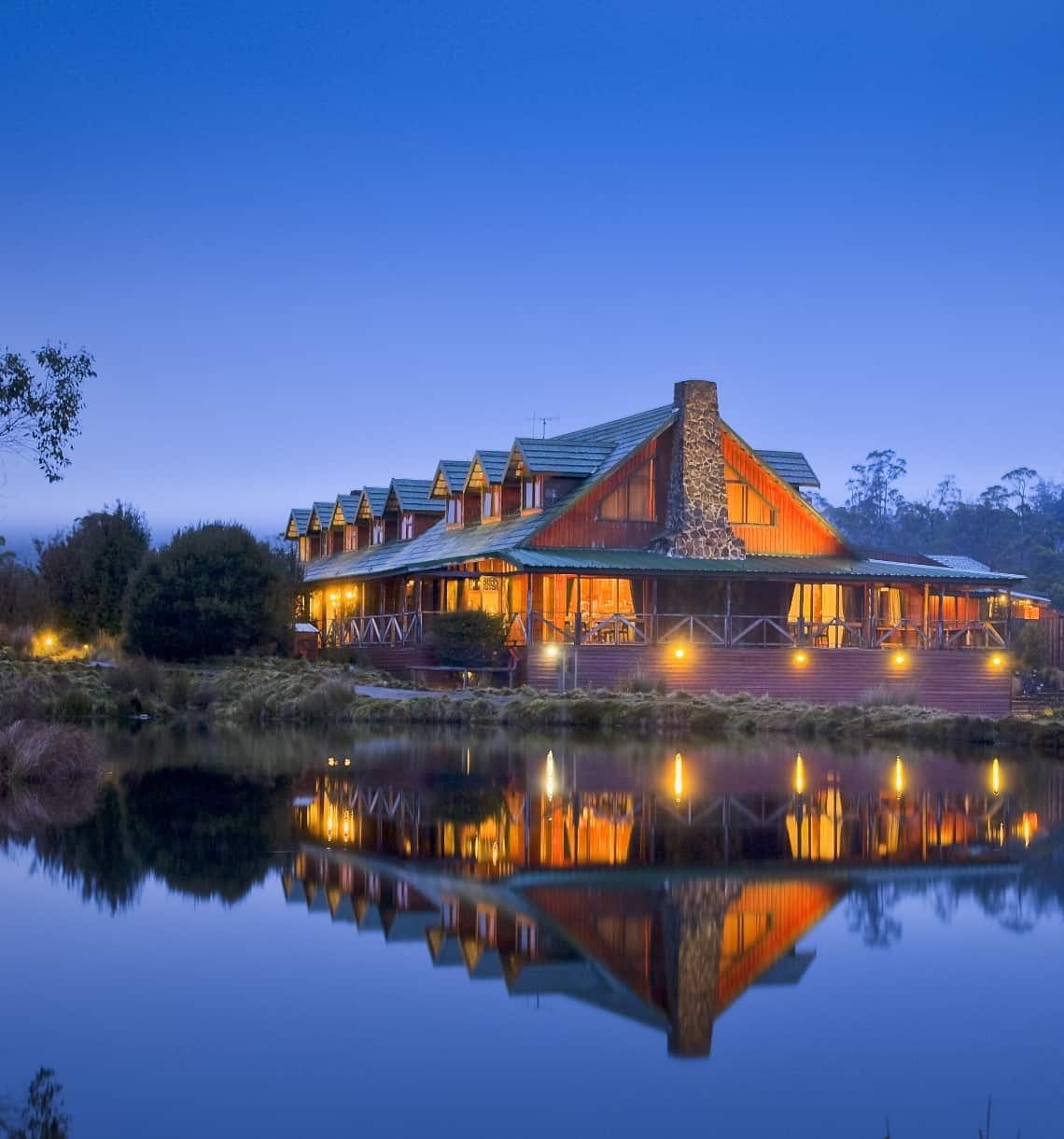 A large rustic house with a stone chimney, lit from inside, reflected in a calm lake at dusk, surrounded by trees and bushes.