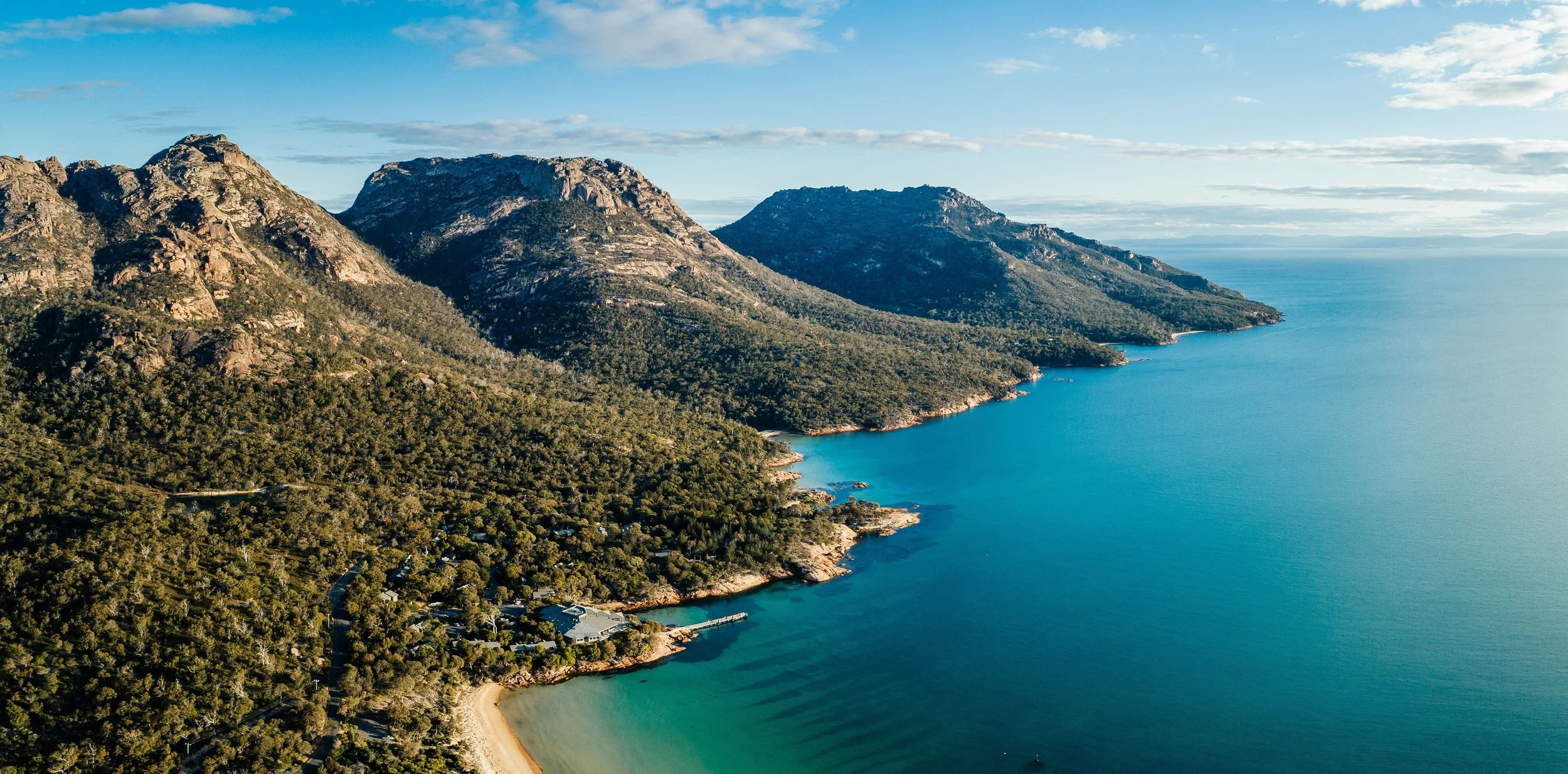 Aerial view of a coastal mountain range with rugged peaks, dense green forest, and a calm blue sea along the shoreline, under a partly cloudy sky.