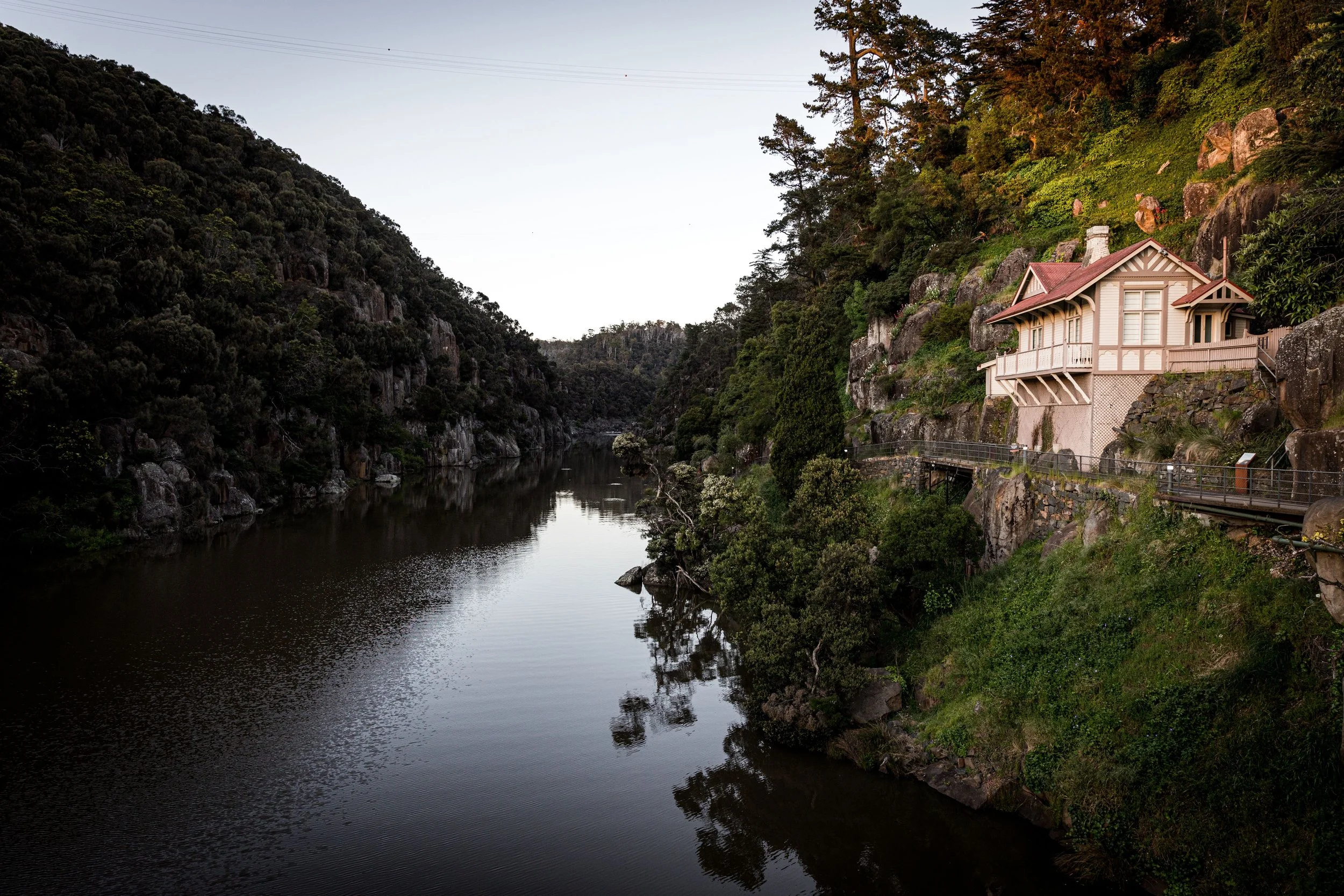 A house built into a rocky hillside next to a river, surrounded by lush greenery and tall trees.