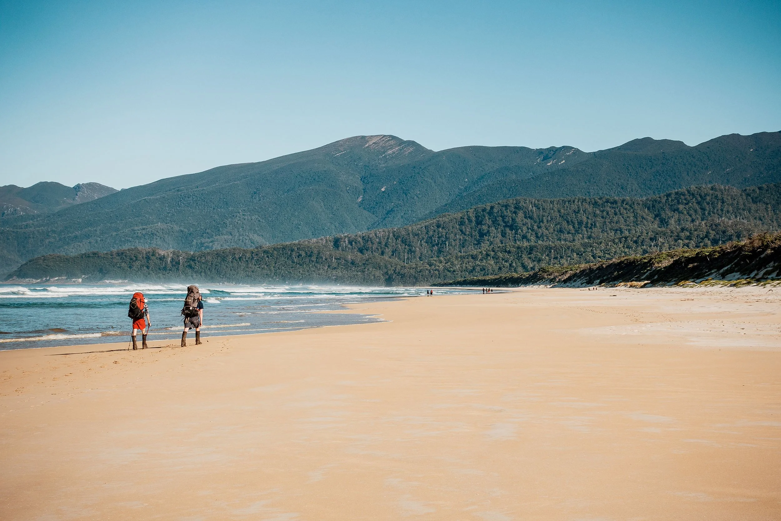 Two hikers with backpacks walking on a sandy beach near the ocean, with green mountains in the background under a clear blue sky.