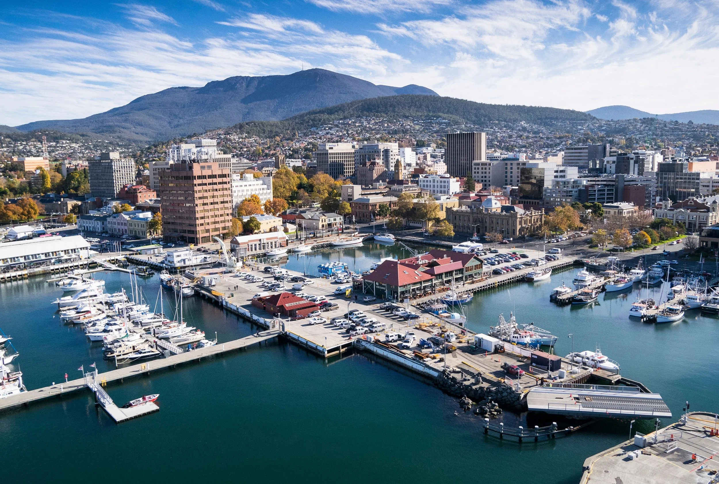 A cityscape with a harbor filled with sailboats and yachts, surrounded by modern buildings, with hills and mountains in the background under a partly cloudy sky.