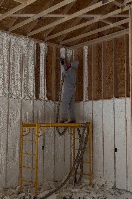 A worker standing on scaffolding applying spray foam insulation between wooden wall studs inside a building under construction.