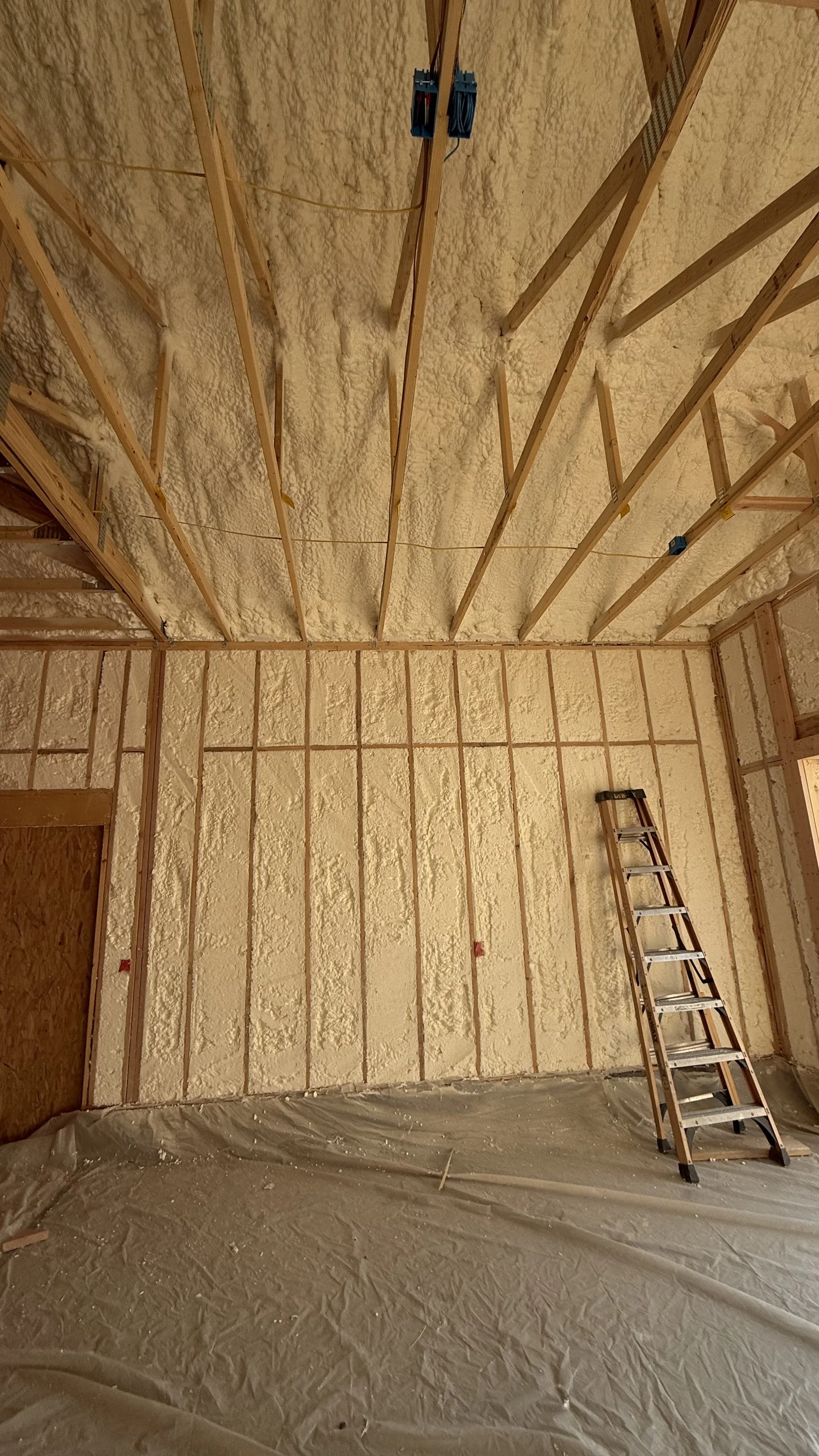 Room under construction with spray foam insulation on walls and ceiling, wooden framing, a ladder leaning against the wall, and plastic sheeting on the floor.