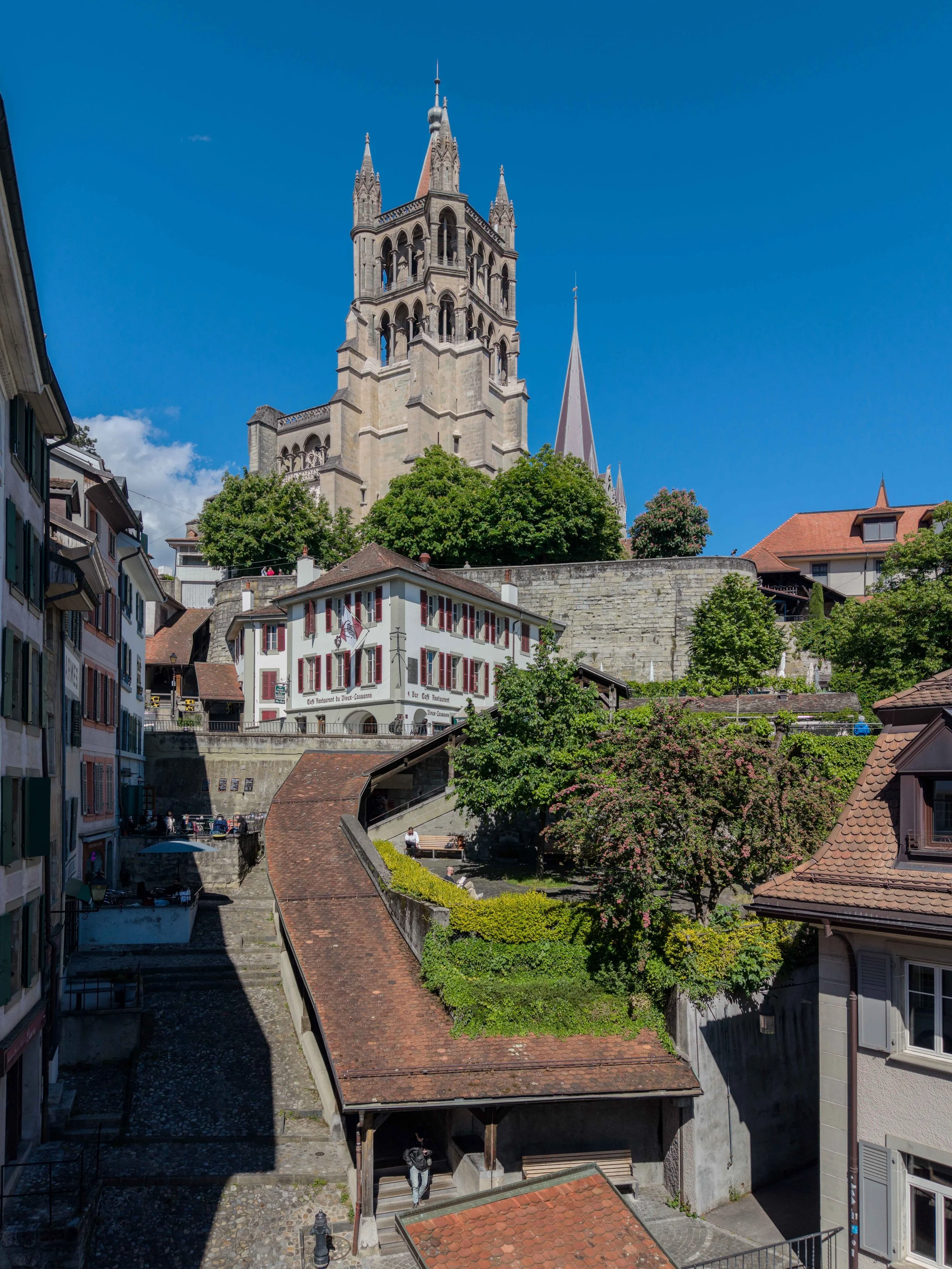 Vue de la vieille ville de Lausanne avec une grande église gothique au fond, entourée de maisons anciennes et d'arbres verts sous un ciel bleu clair.