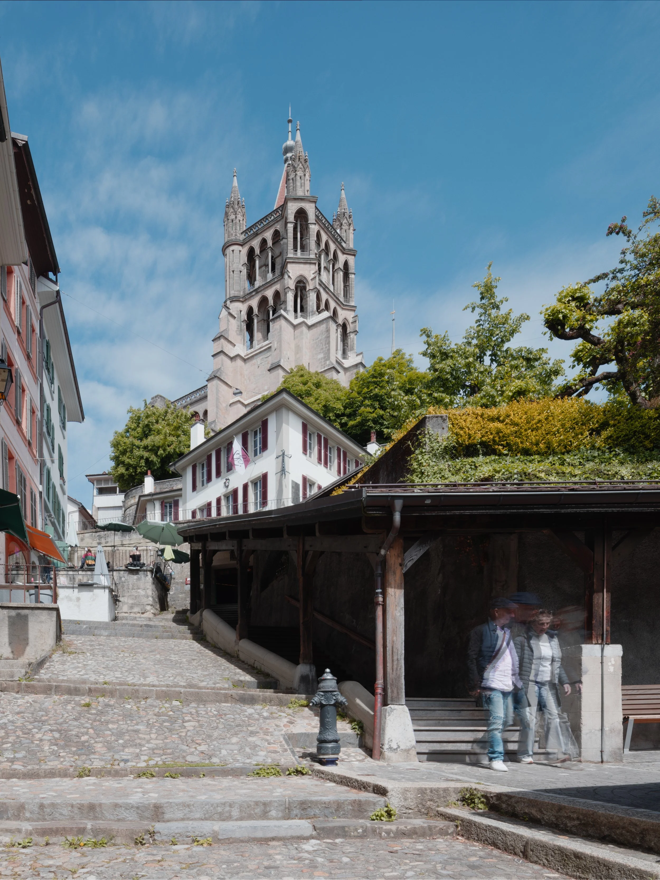 Une scène urbaine avec un bâtiment en pierre avec des tours pointues, et des bâtiments colorés en premier plan, des arbres verts, un ciel bleu avec quelques nuages et des personnes marchant sous une petite structure en bois avec un toit.