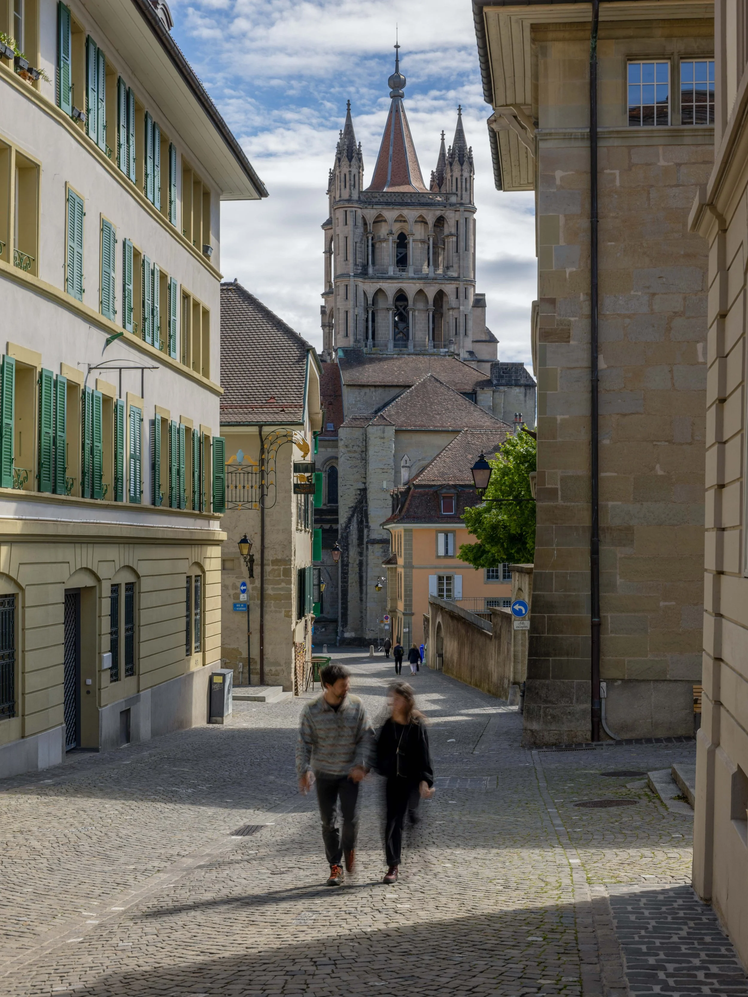 Une rue pavée avec deux personnes qui marchent, entourée de bâtiments anciens avec des fenêtres à volets verts, et une tour d'église en arrière-plan sous un ciel partiellement nuageux.