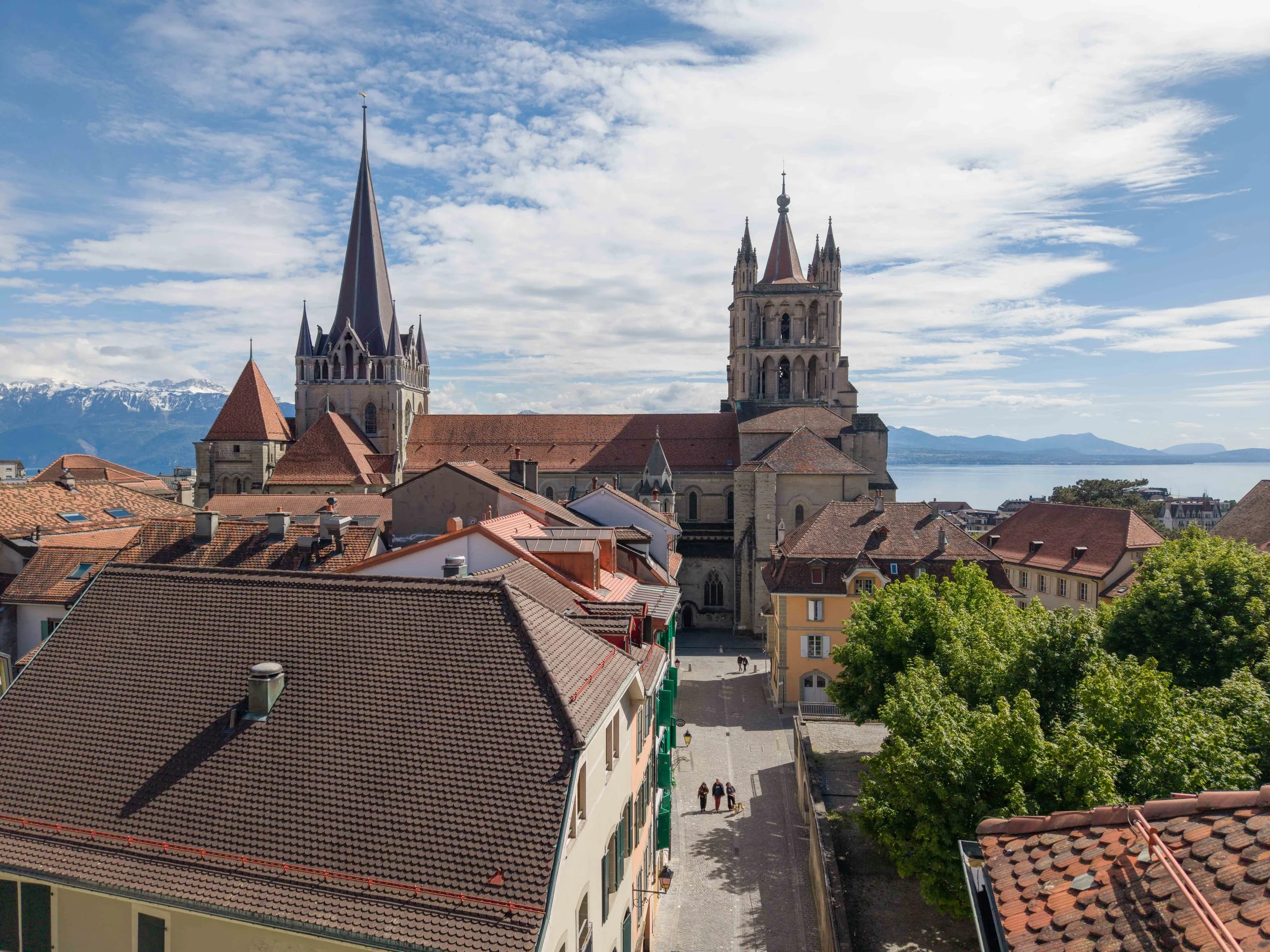 Vue d'ensemble de toits en tuiles rouges de bâtiments dans une ville avec deux églises gothiques et un lac en arrière-plan, montagnes enneigées au loin, ciel partiellement nuageux.